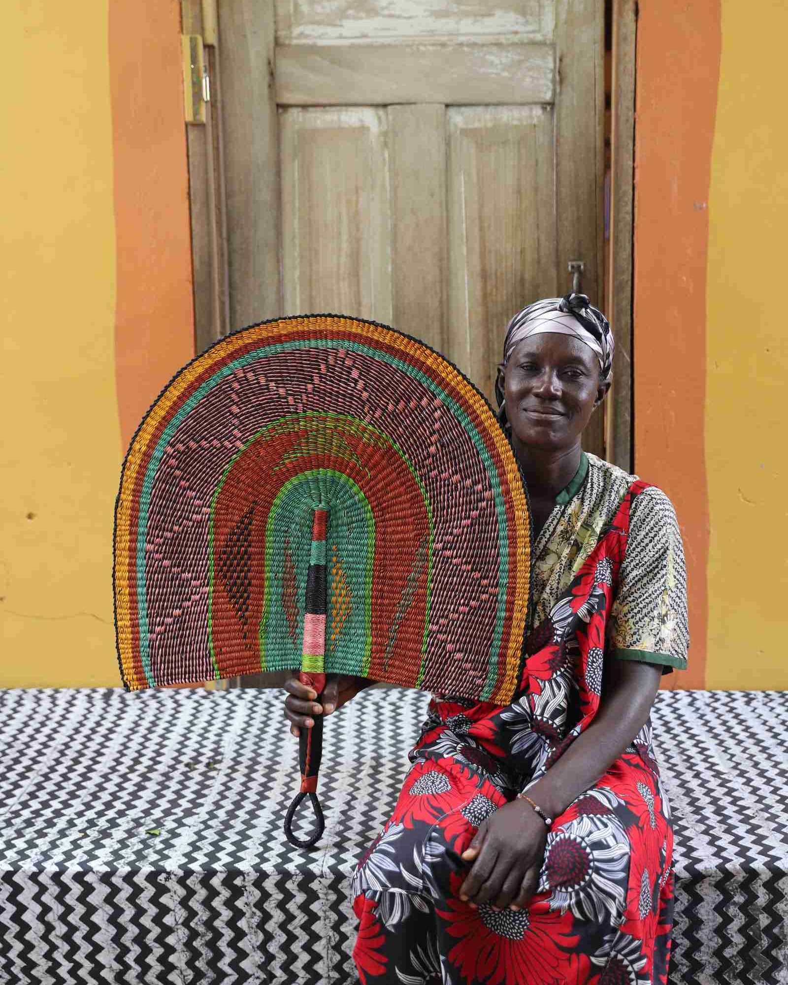 Naba Penga wall fan in natural straw with green, orange and black concentric bands, held by a seated person for scale.