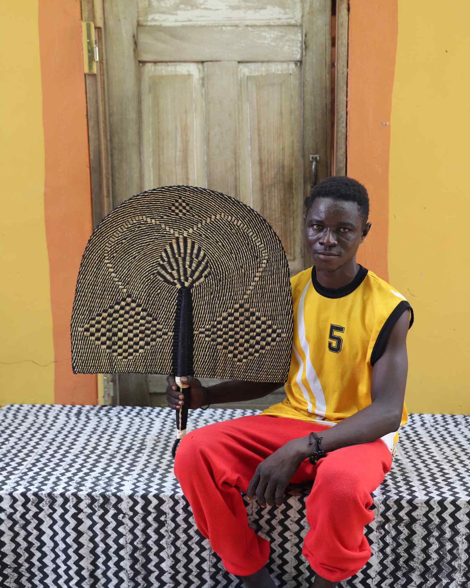 Weaver holding a Naba Penga handwoven wall fan in natural straw and black geometric check, in Bolgatanga, Ghana.