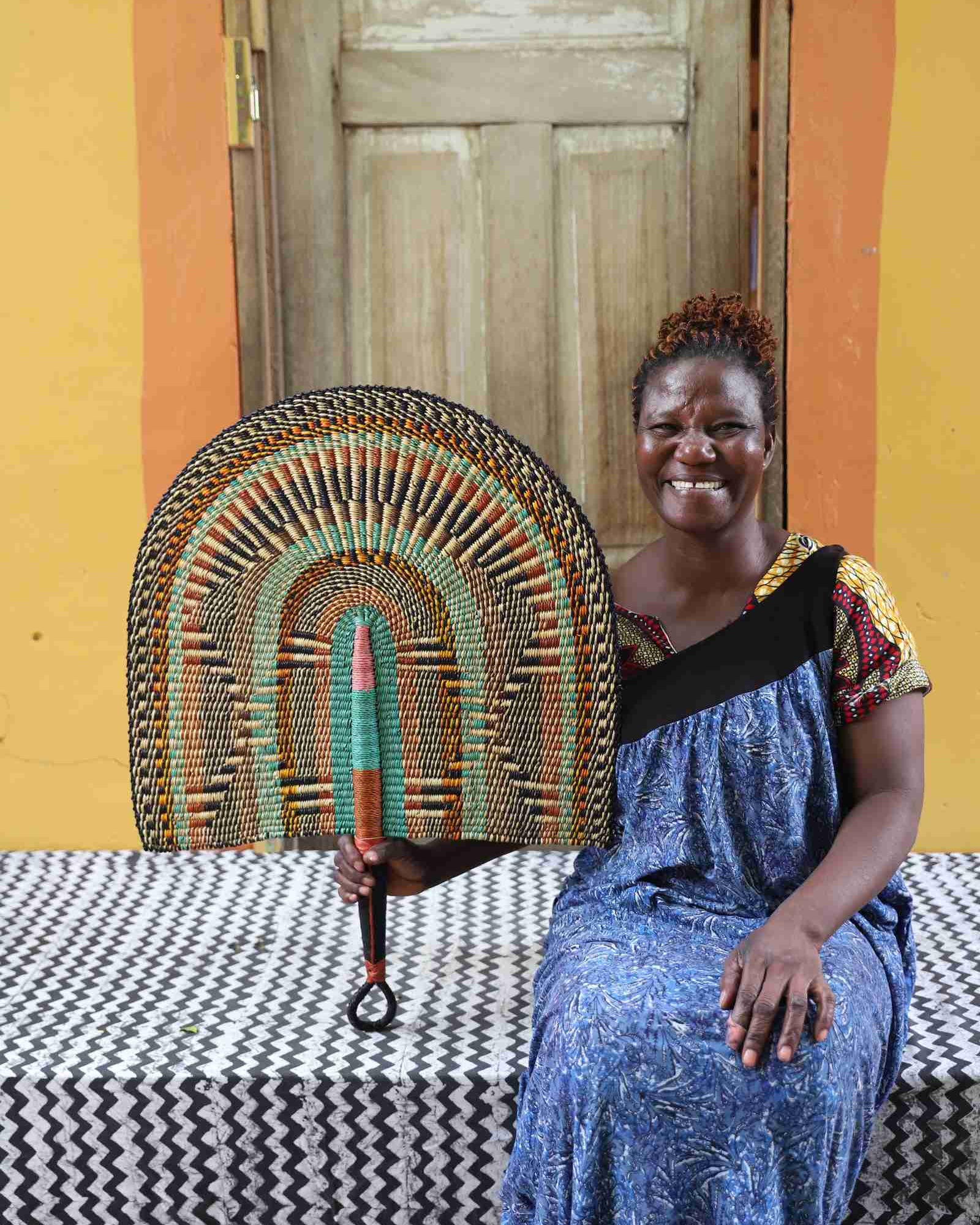 Naba Penga wall fan in natural straw with rust, green and teal concentric arches, held by a seated person for scale.