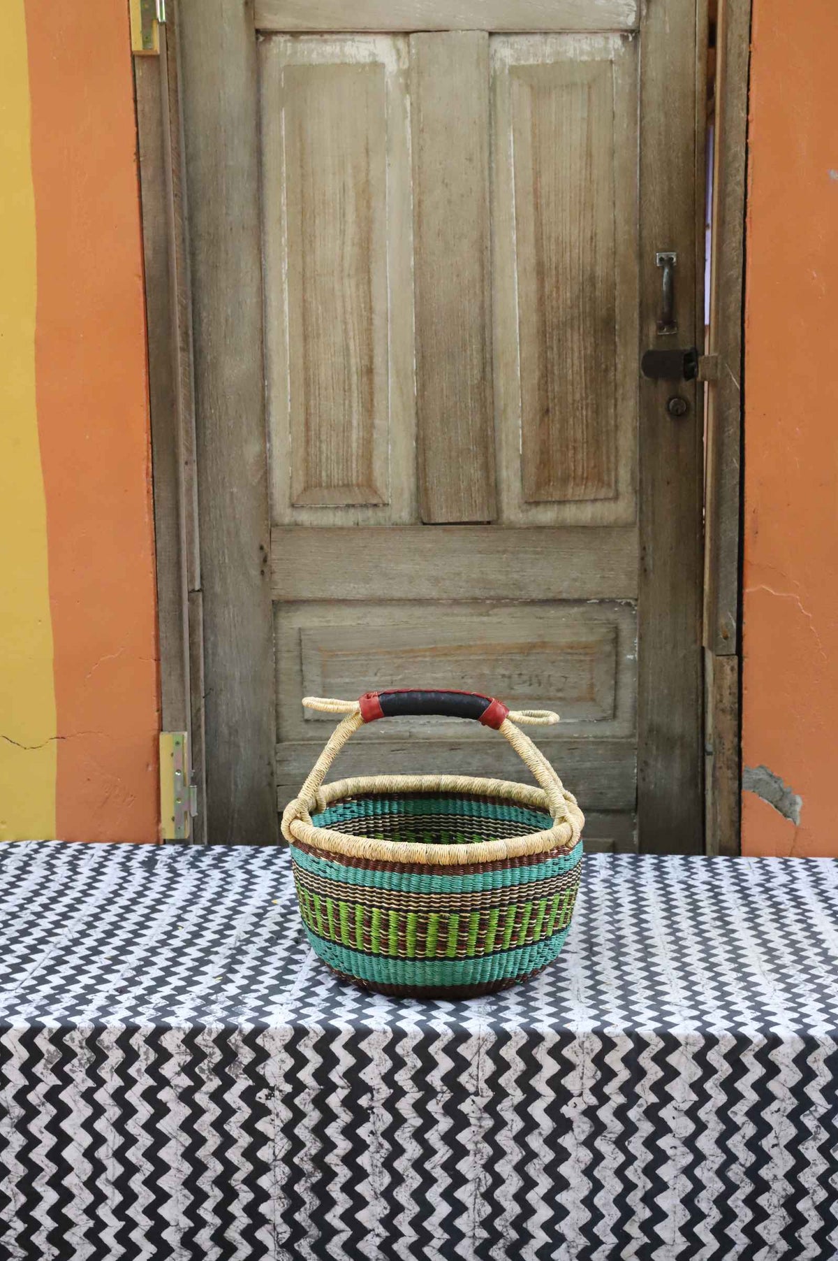 Handwoven round Bolga shopping basket in natural straw with green and teal woven bands and a leather-wrapped handle.