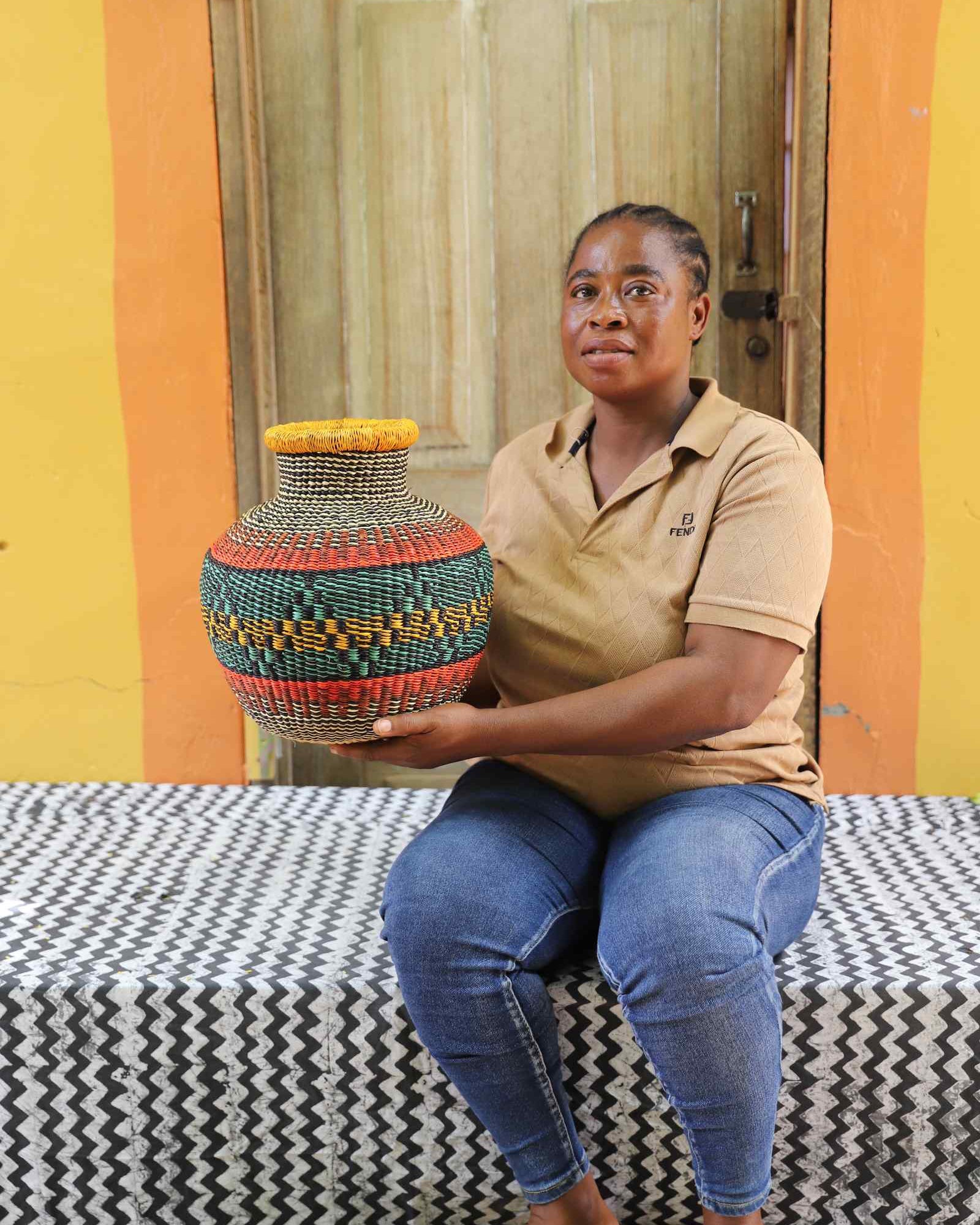 Small round Bolga pot basket in teal, orange and yellow diamond weave, held by a seated woman for scale.