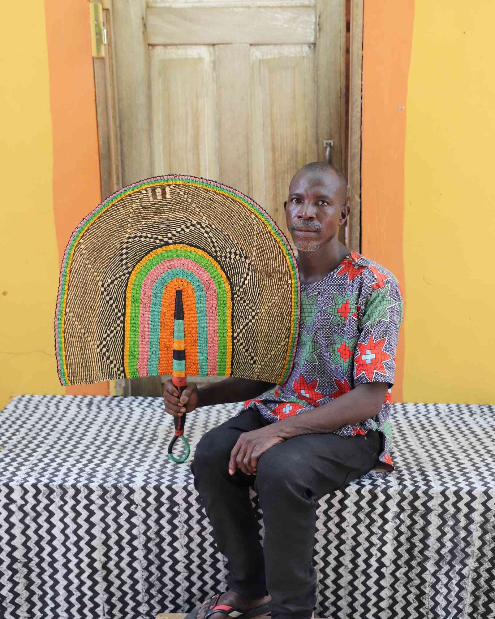 Weaver holding a Naba Penga handwoven wall fan in natural straw with pink, green and orange inner arches, in Bolgatanga.