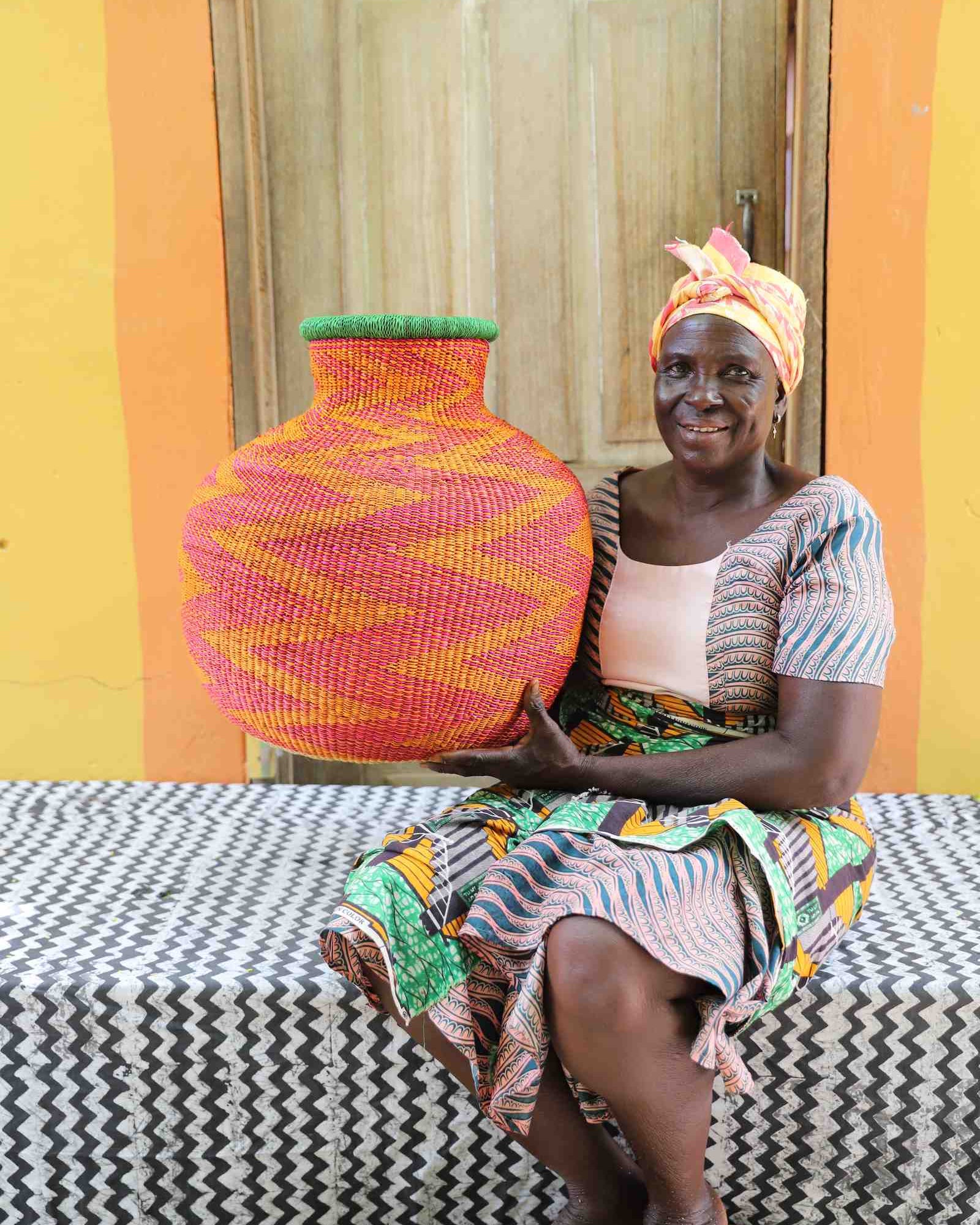 Large round Bolga pot basket in pink and yellow zigzag weave with a green rim, held by a seated woman for scale.