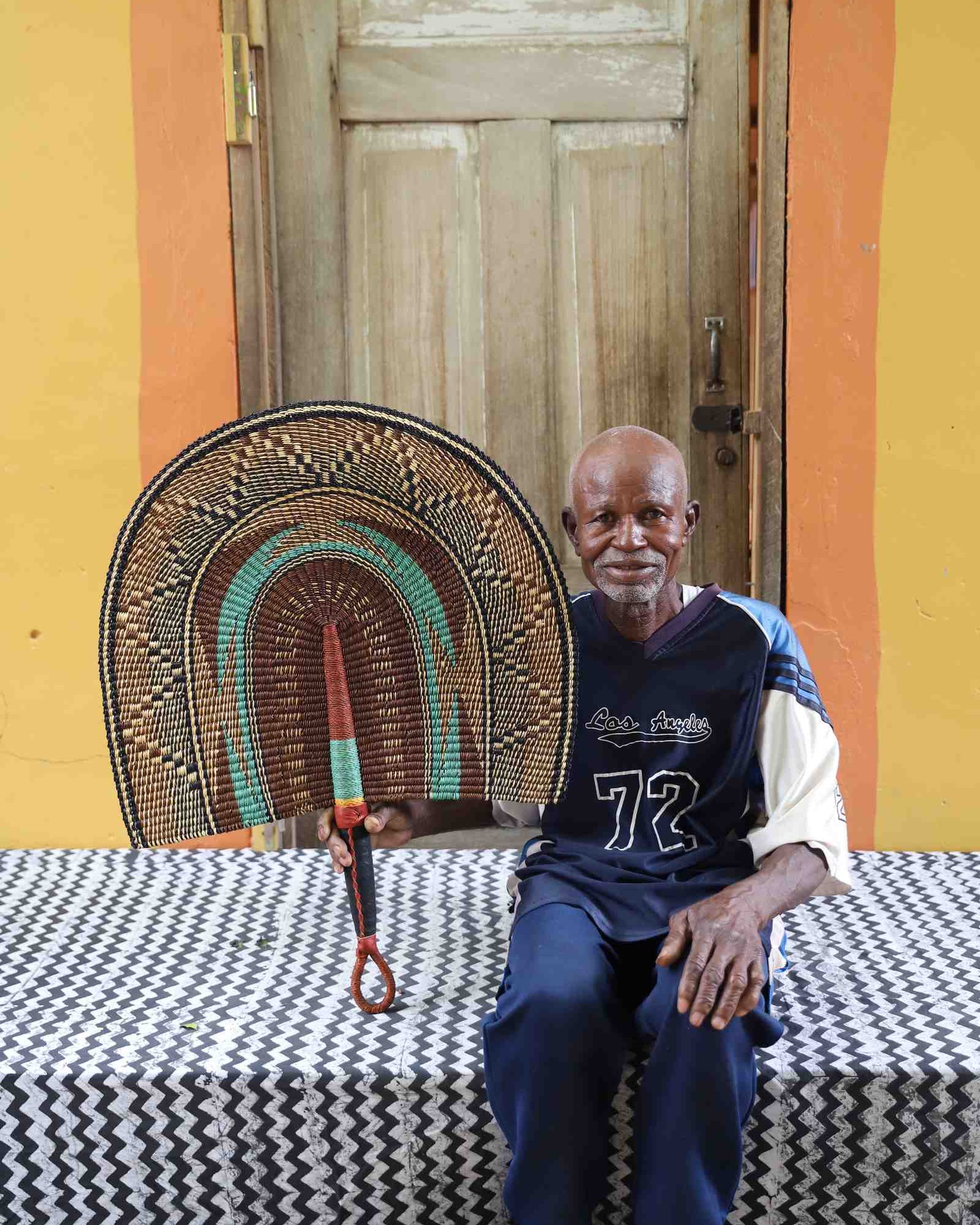 Naba Penga wall fan in natural straw with a teal arch and orange patterned border, held by a seated person for scale.
