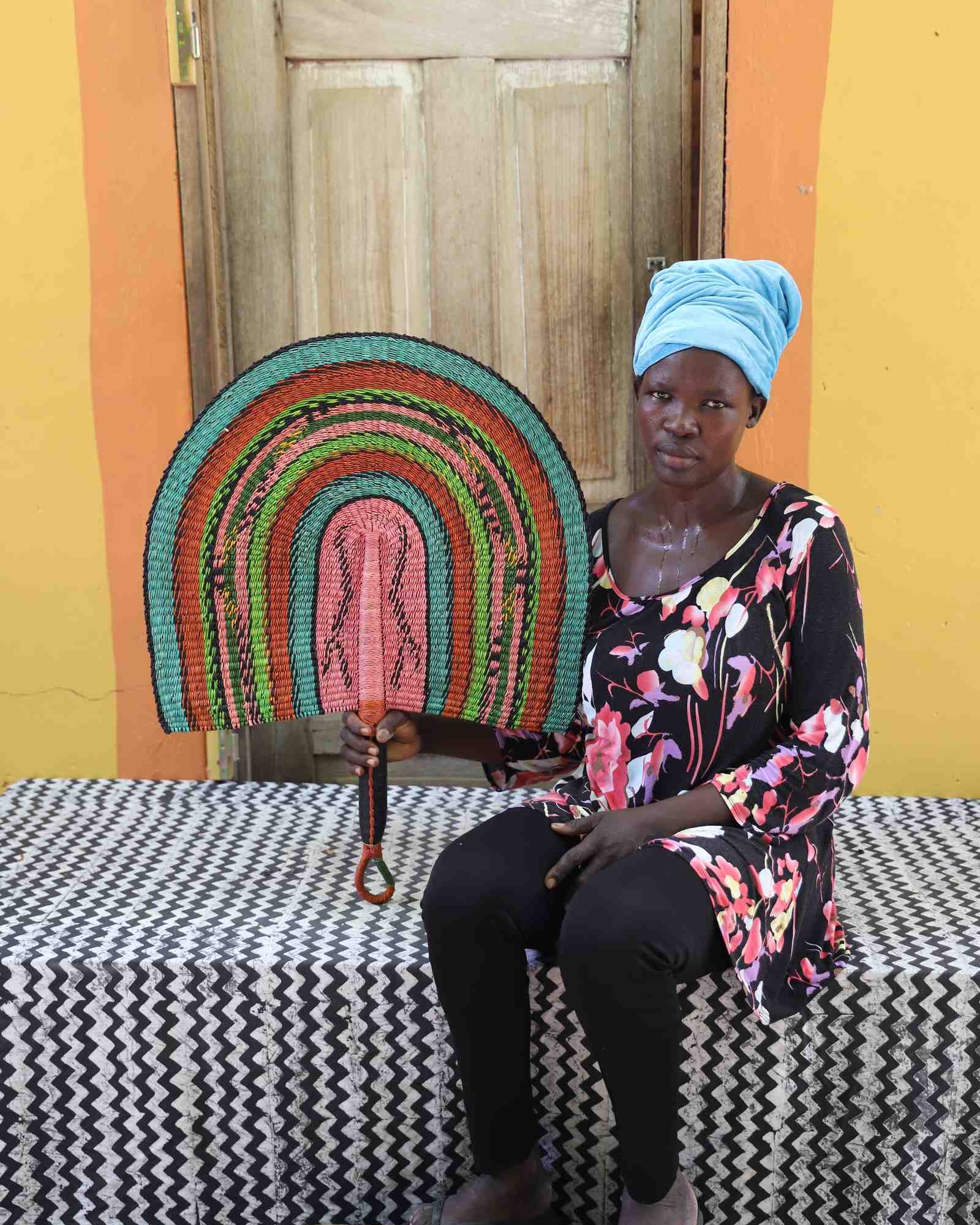 Weaver holding a Naba Penga handwoven wall fan in teal, terracotta and pink, in Bolgatanga, Ghana.