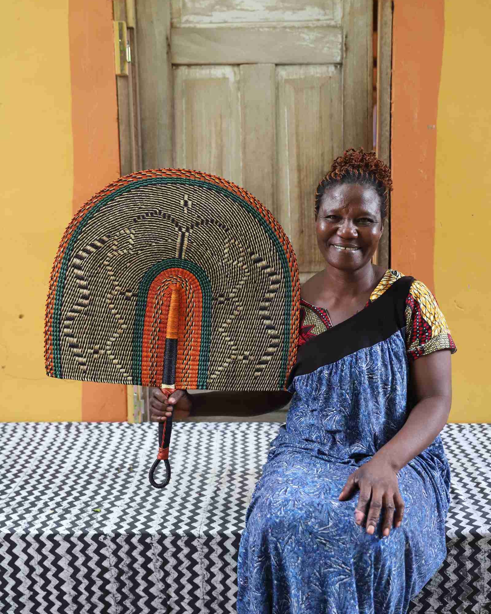 Naba Penga wall fan in natural straw with an orange and green banded border, held by a seated person for scale.