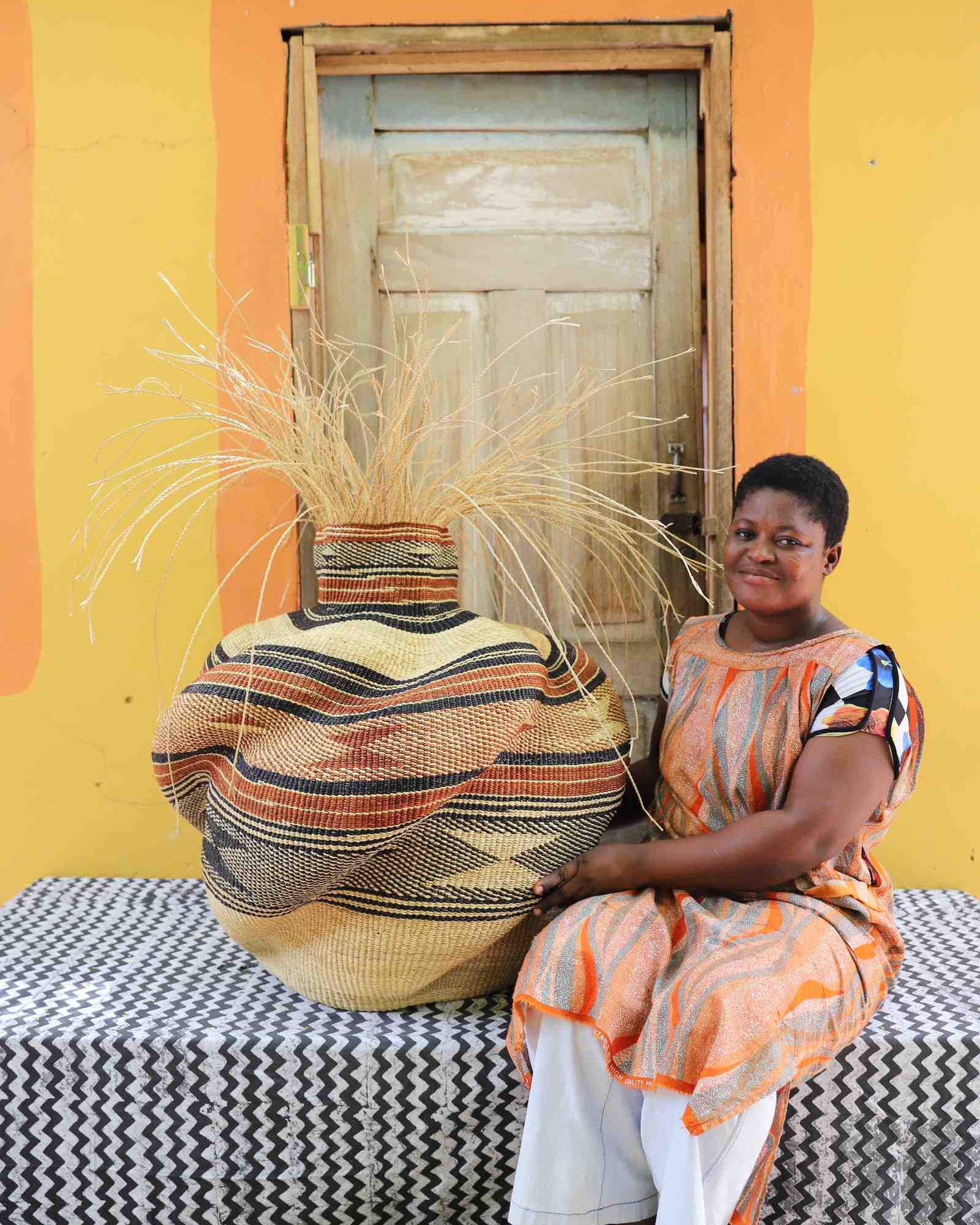 Round Bolga basket in tan with a rust and black striped band and loose straw fronds, shown beside a seated woman for scale.