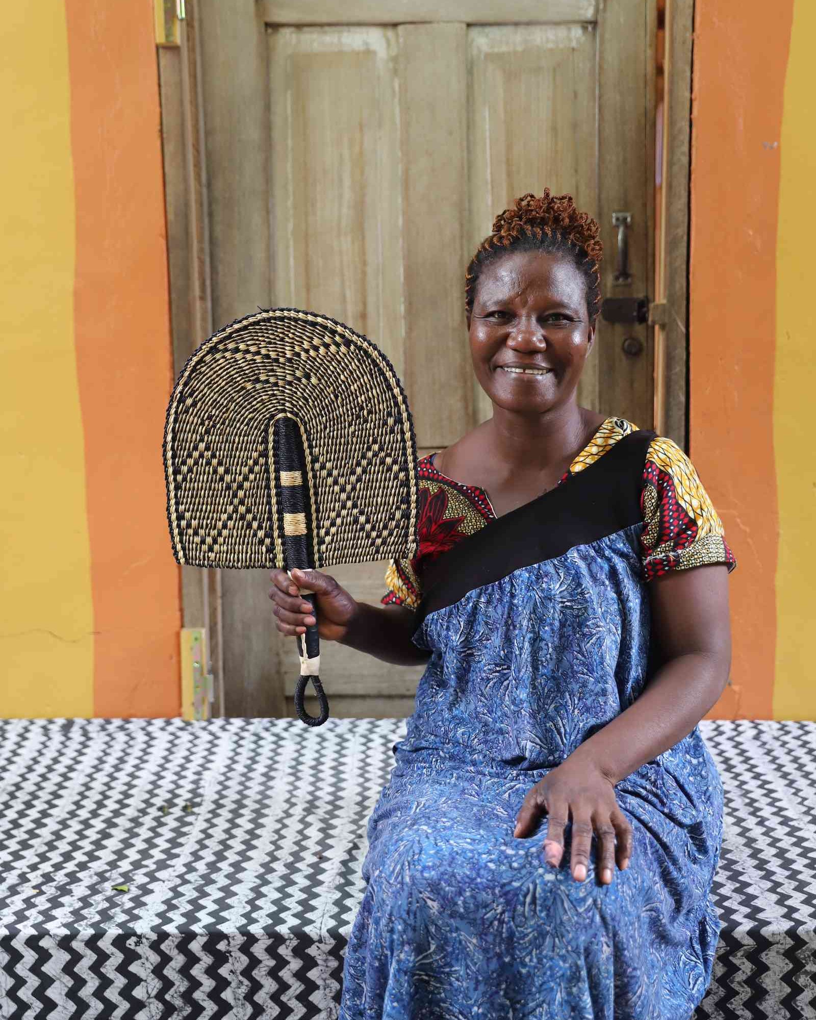 Bolga hand fan in natural straw with a dark chevron pattern, held by a seated person for scale.