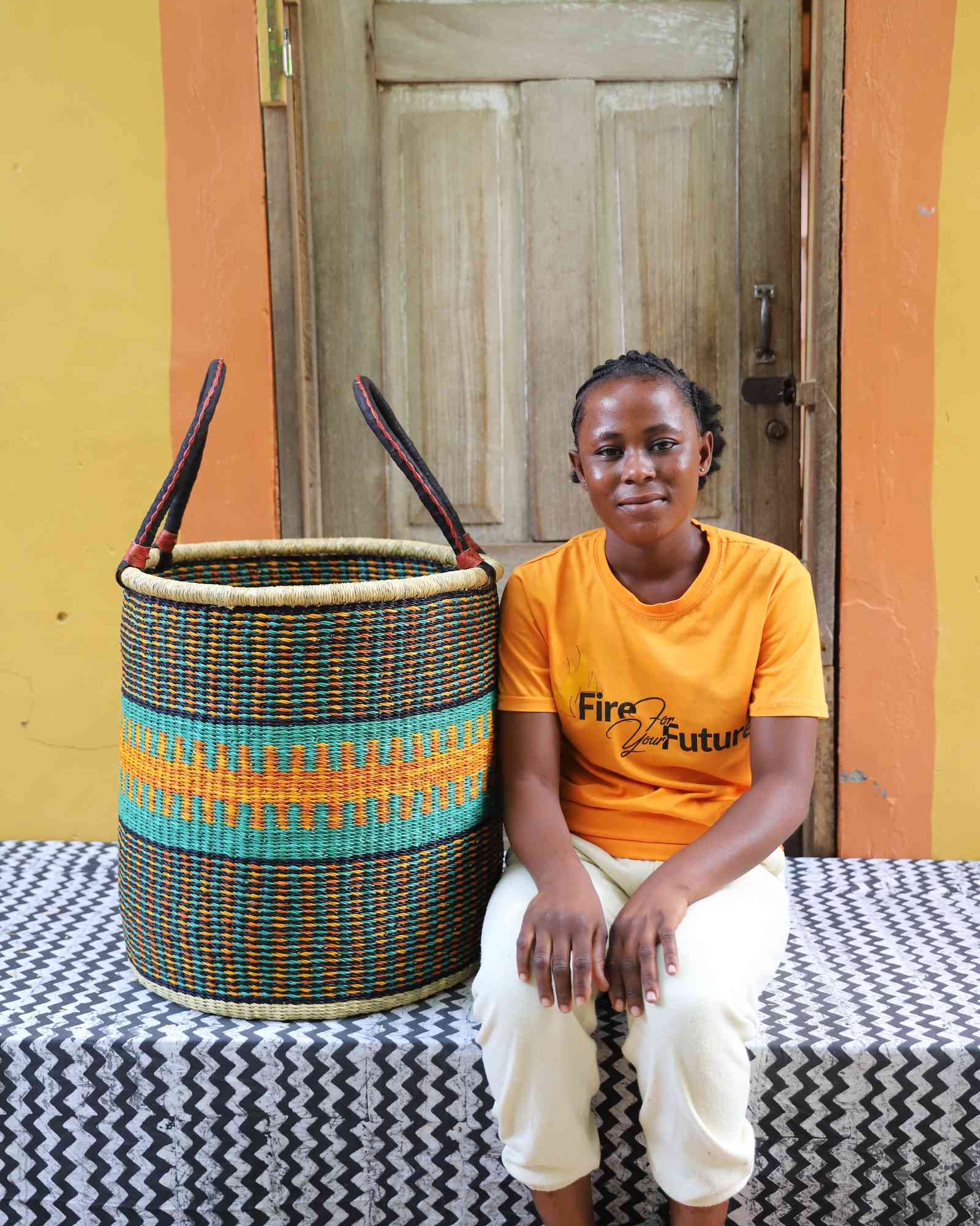 Tall open Bolga laundry basket with a teal and orange patterned band, shown beside a seated woman for scale.