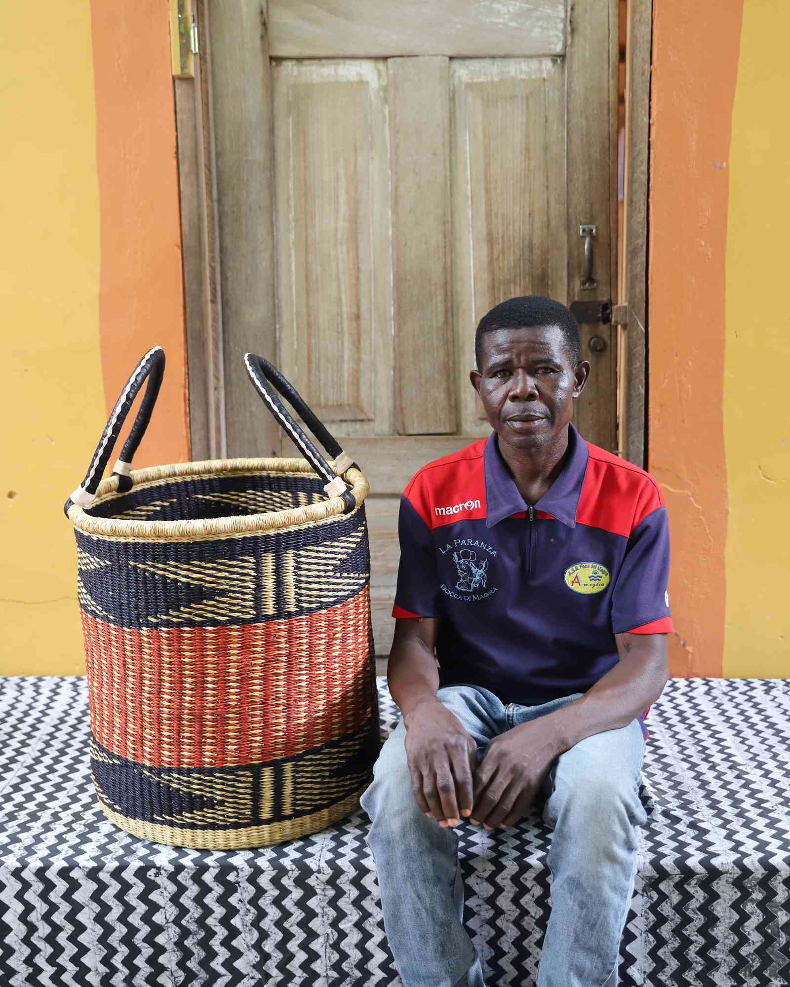 Open laundry basket in natural straw with an orange and black zigzag band, shown beside a seated person for scale.
