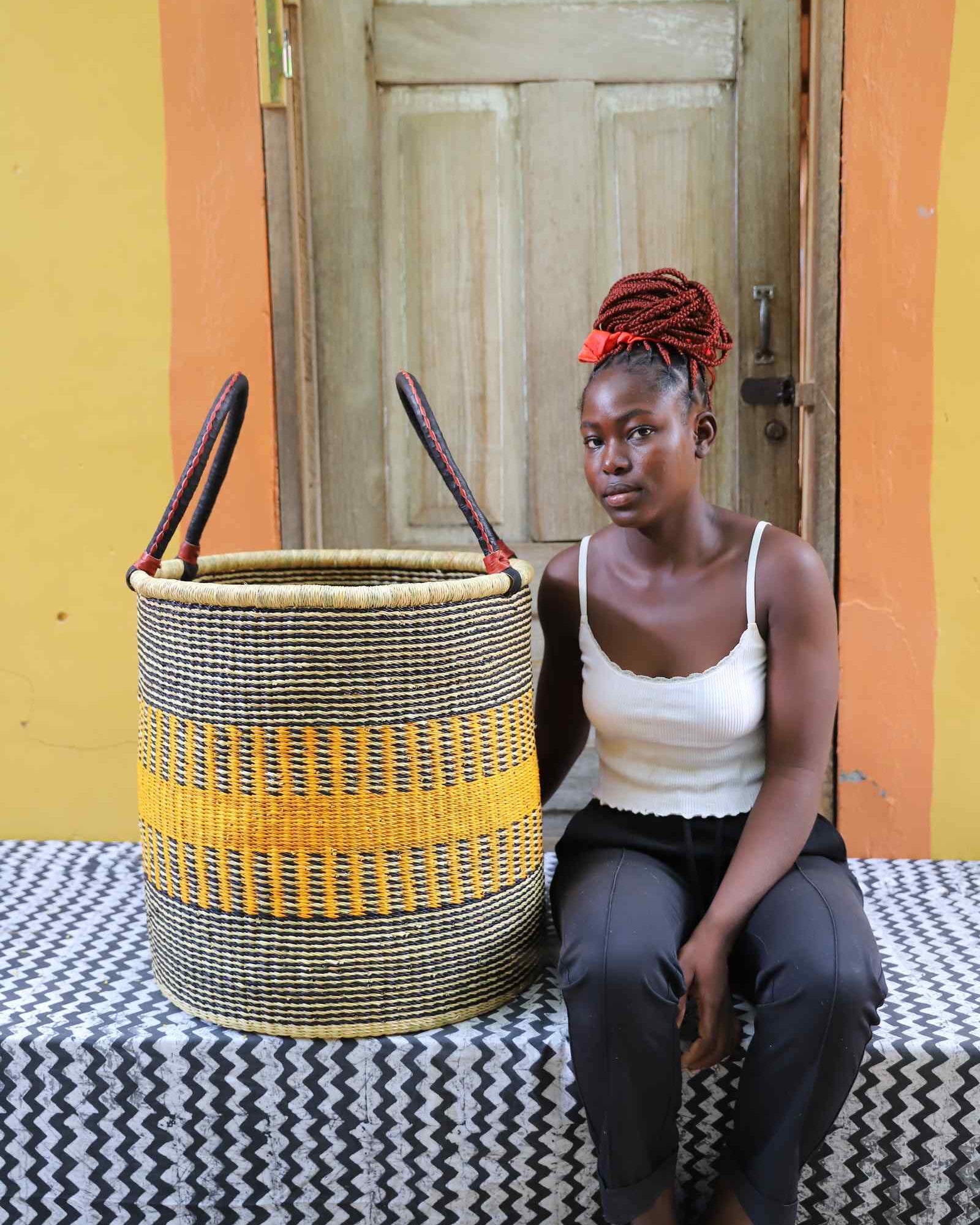 Tall open Bolga laundry basket with a bold yellow stripe band, shown beside a seated woman for scale.