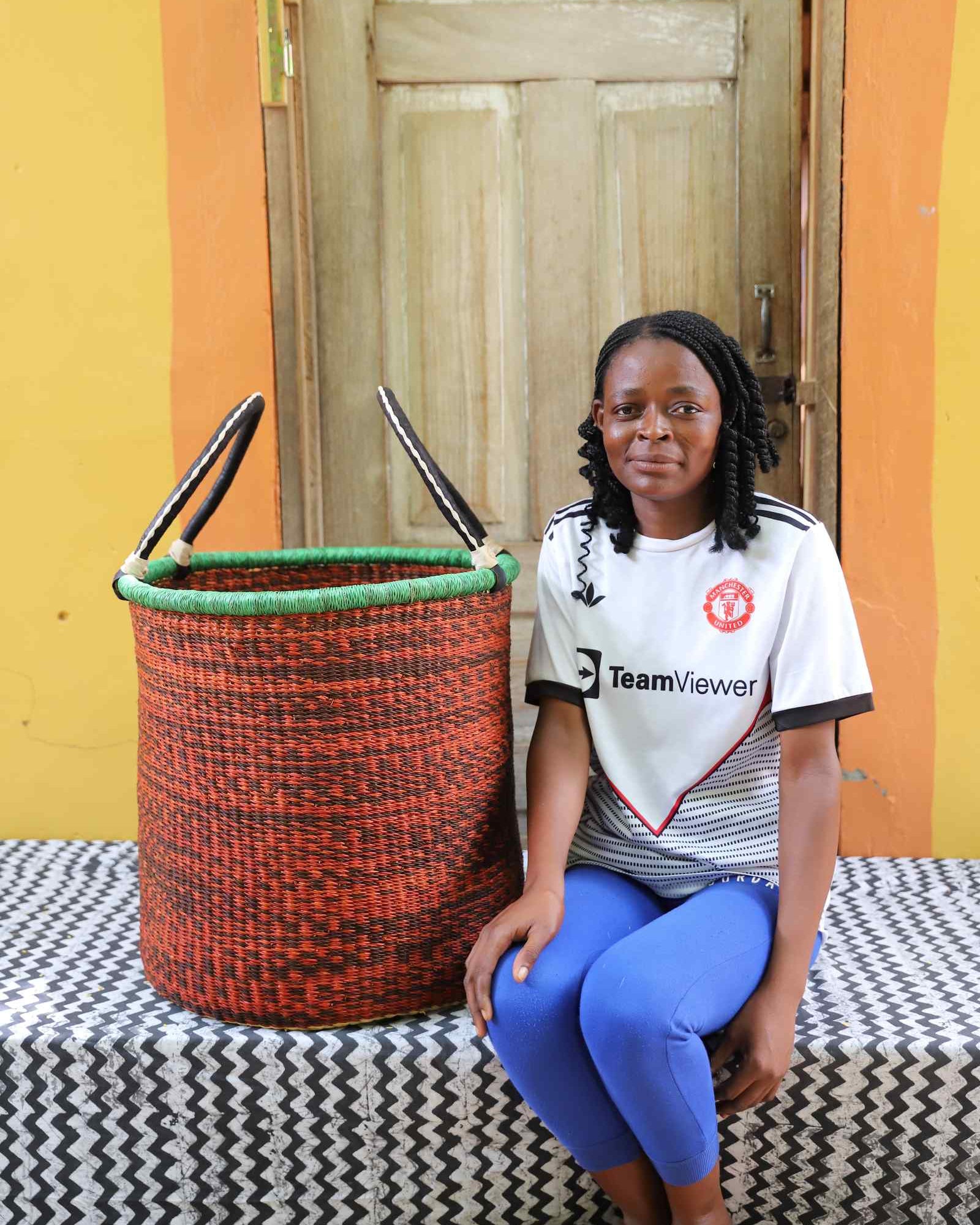 Tall open Bolga laundry basket in speckled rust-red weave, shown beside a seated woman for scale.