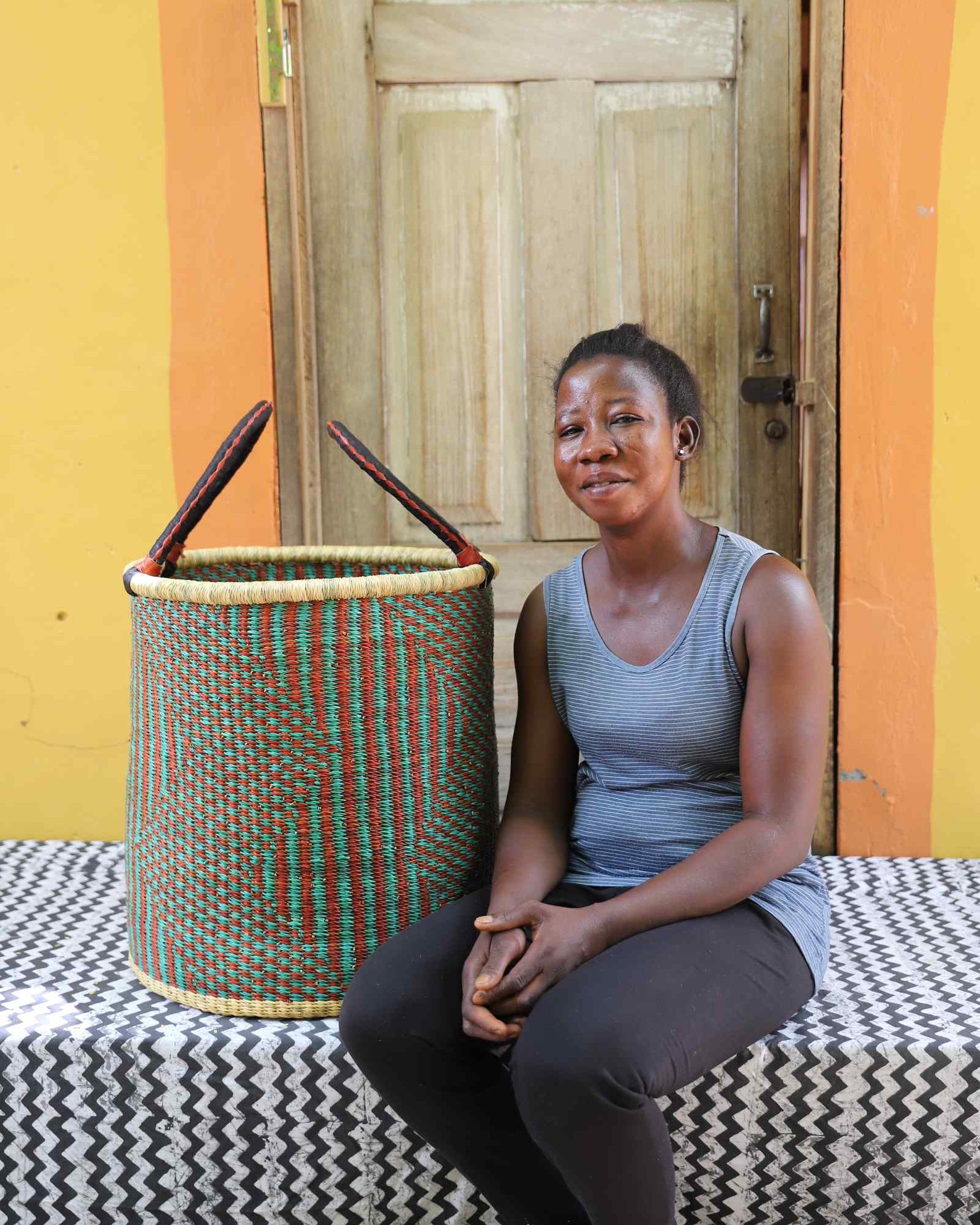 Tall open Bolga laundry basket in a green and rust striped weave, shown beside a seated woman for scale.