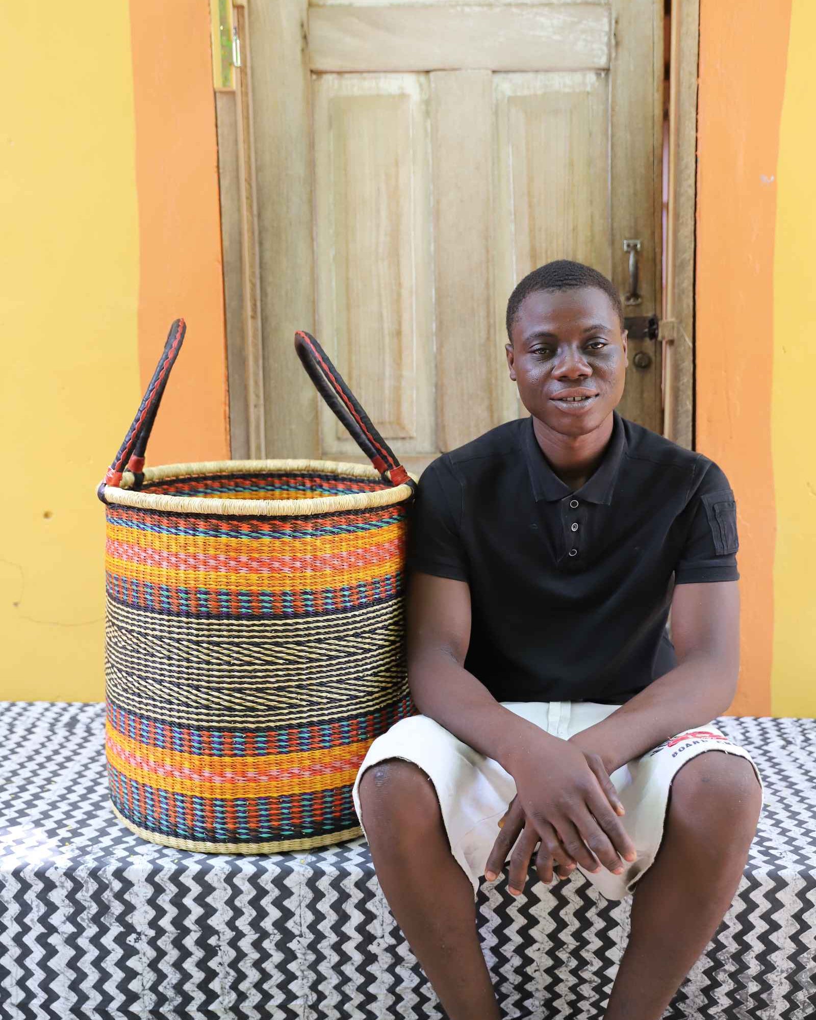 Tall open Bolga laundry basket with an orange, blue and cream striped band, shown beside a seated man for scale.