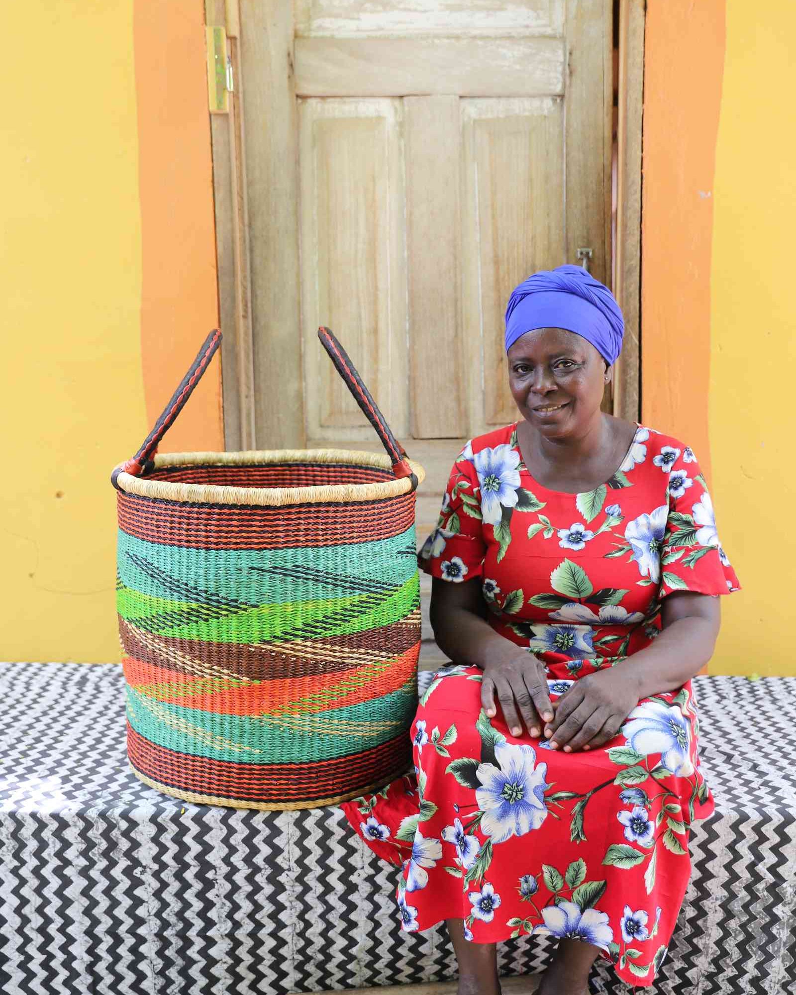 Tall open Bolga laundry basket with a multicolour diagonal striped weave, shown beside a seated woman for scale.
