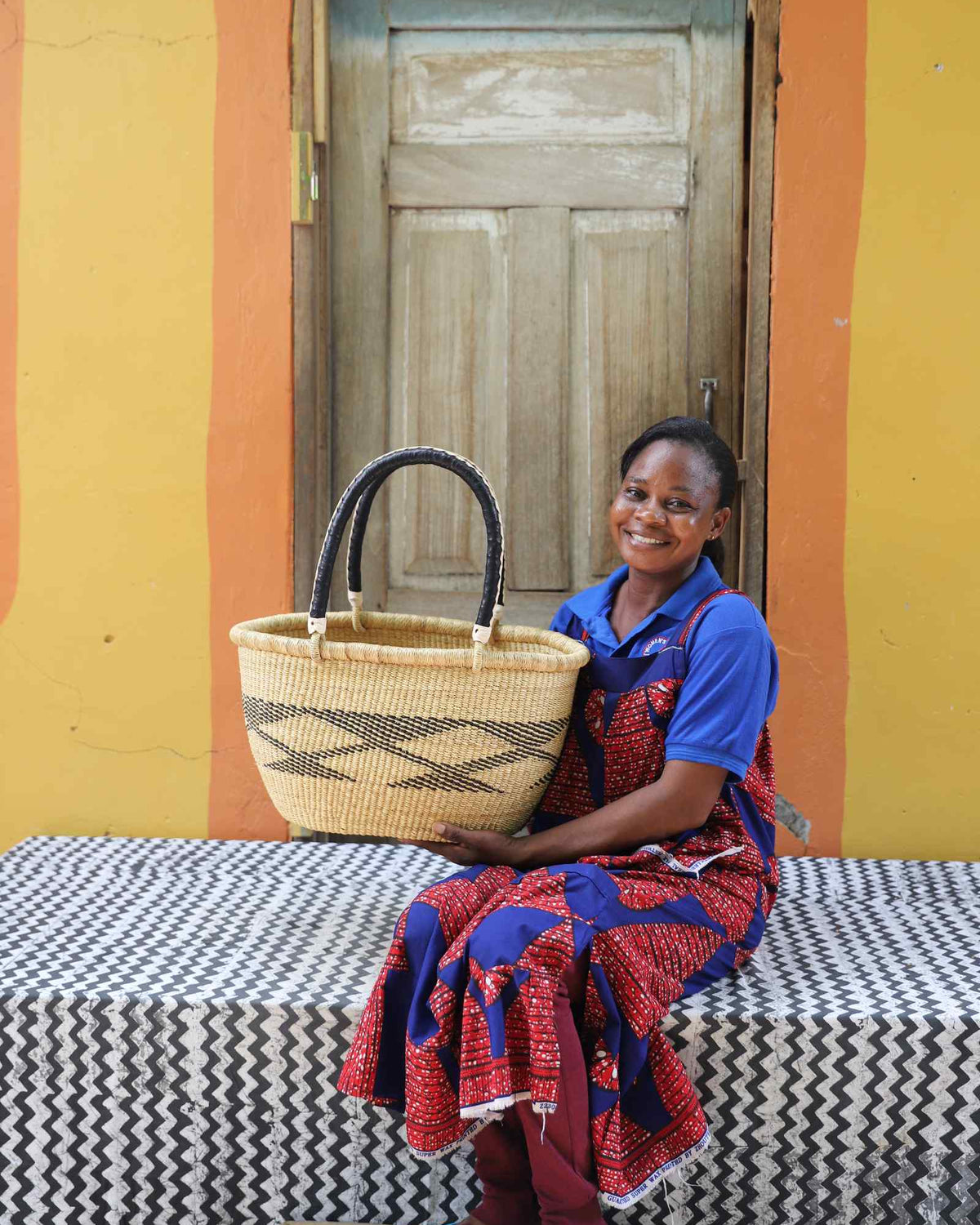 Medium oval Bolga shopping basket in natural with dark bands, held by a seated woman in a printed dress.
