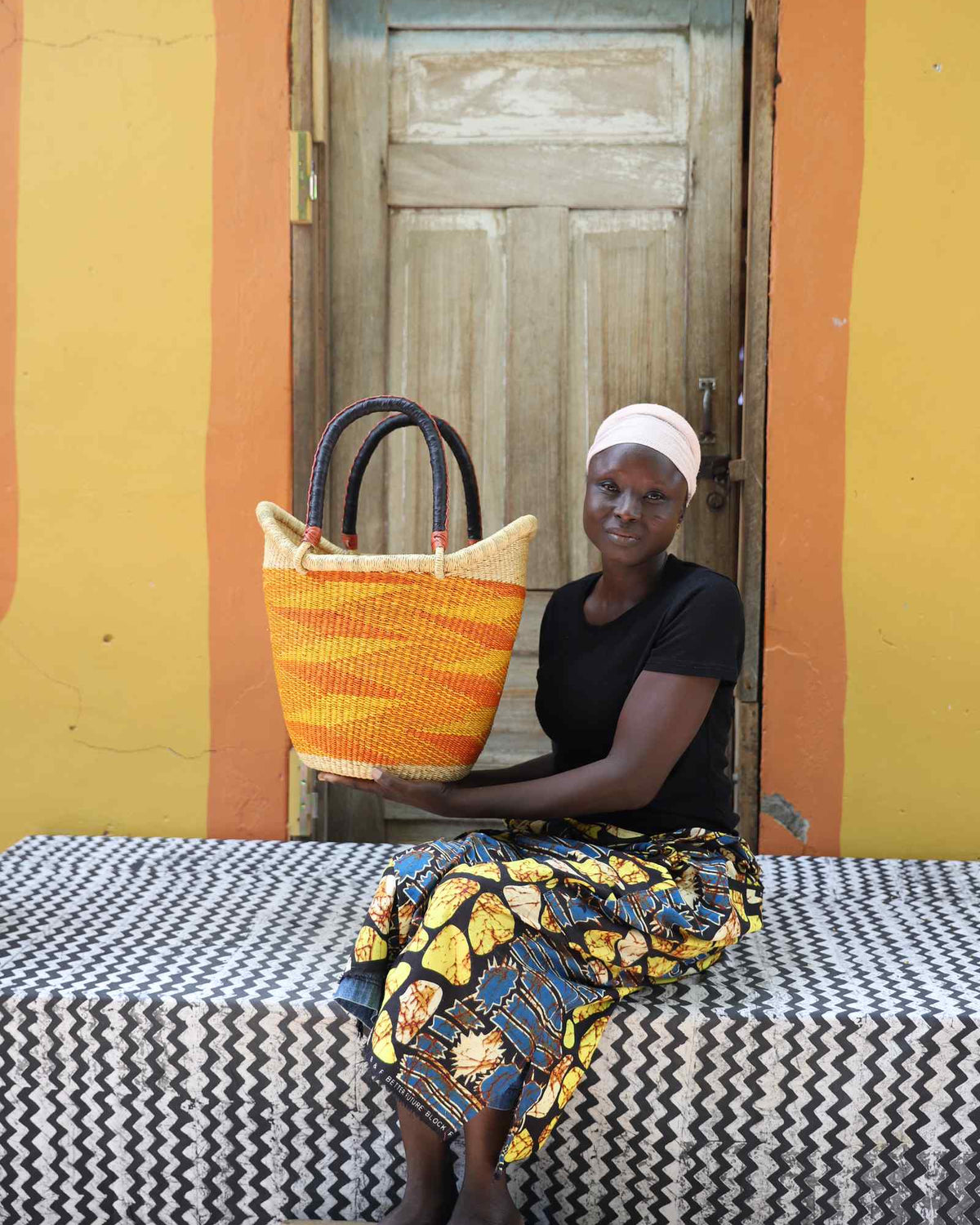 Medium Nyariga Bolga shopping basket in dyed bold orange, held by a seated woman in a floral skirt.
