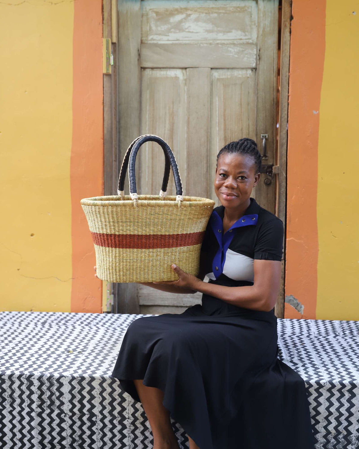 Large Bolga Zaane Tia market basket with rust-orange band, held by a seated woman in a black dress.