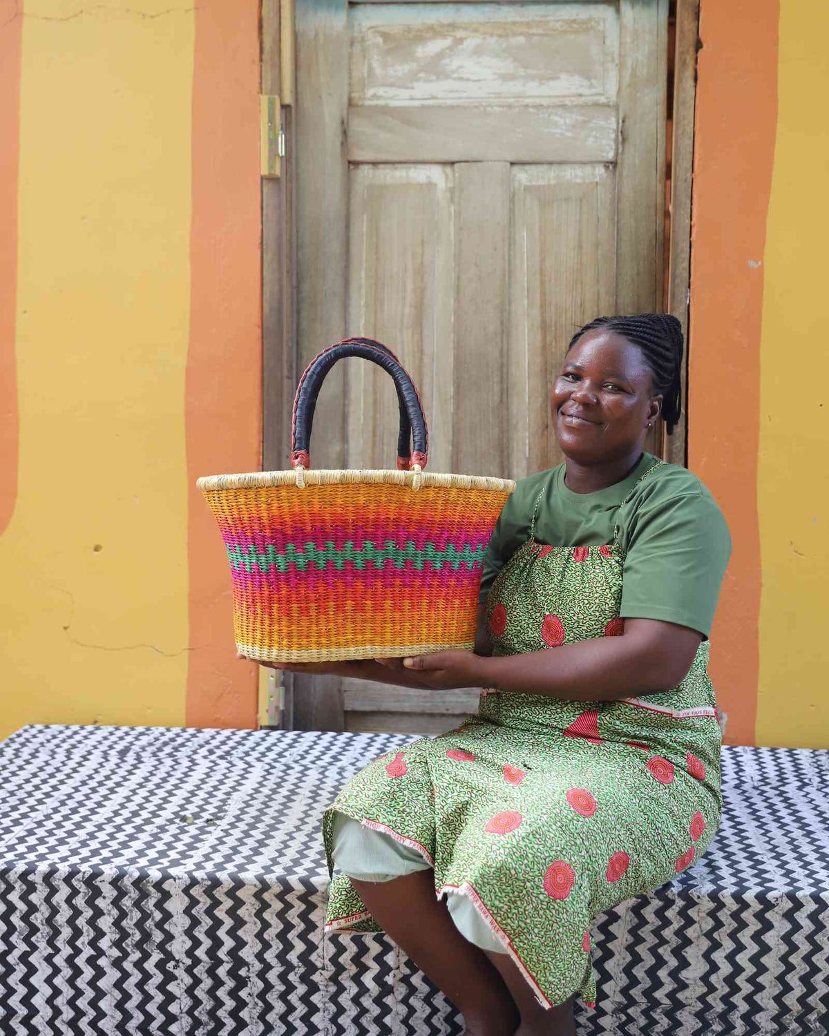 Large Bolga Zaane Tia market basket in multicolour stripes, held by a seated woman in green.