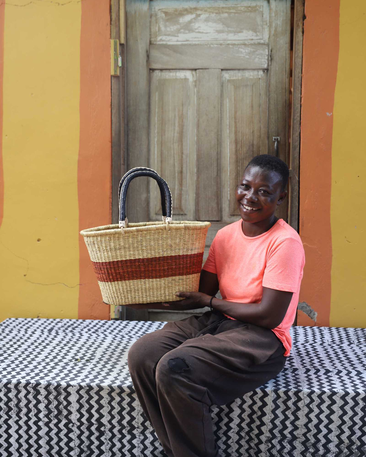 Medium Bolga Zaane Tia market basket with rust-orange band, held by a seated woman in coral.
