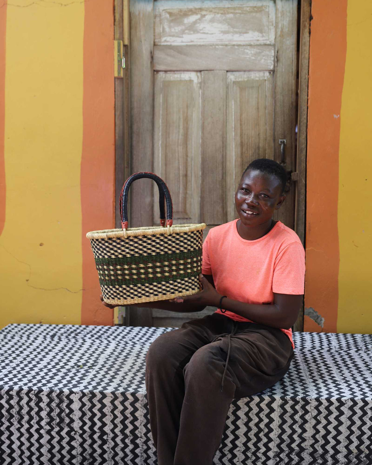 Small Bolga Zaane Tia market basket with dark and orange zigzag band, held by a seated woman in yellow.