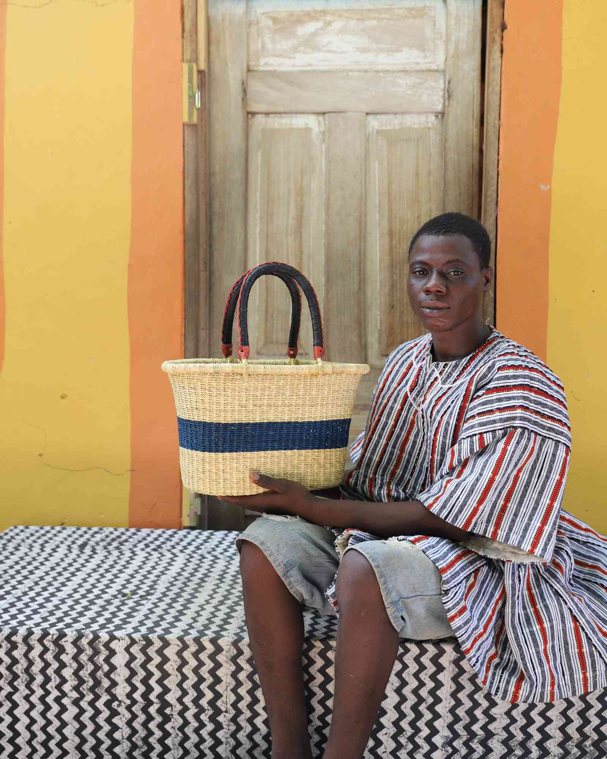 Small Bolga Zaane Tia market basket with navy band, held by a seated man in a striped tunic.