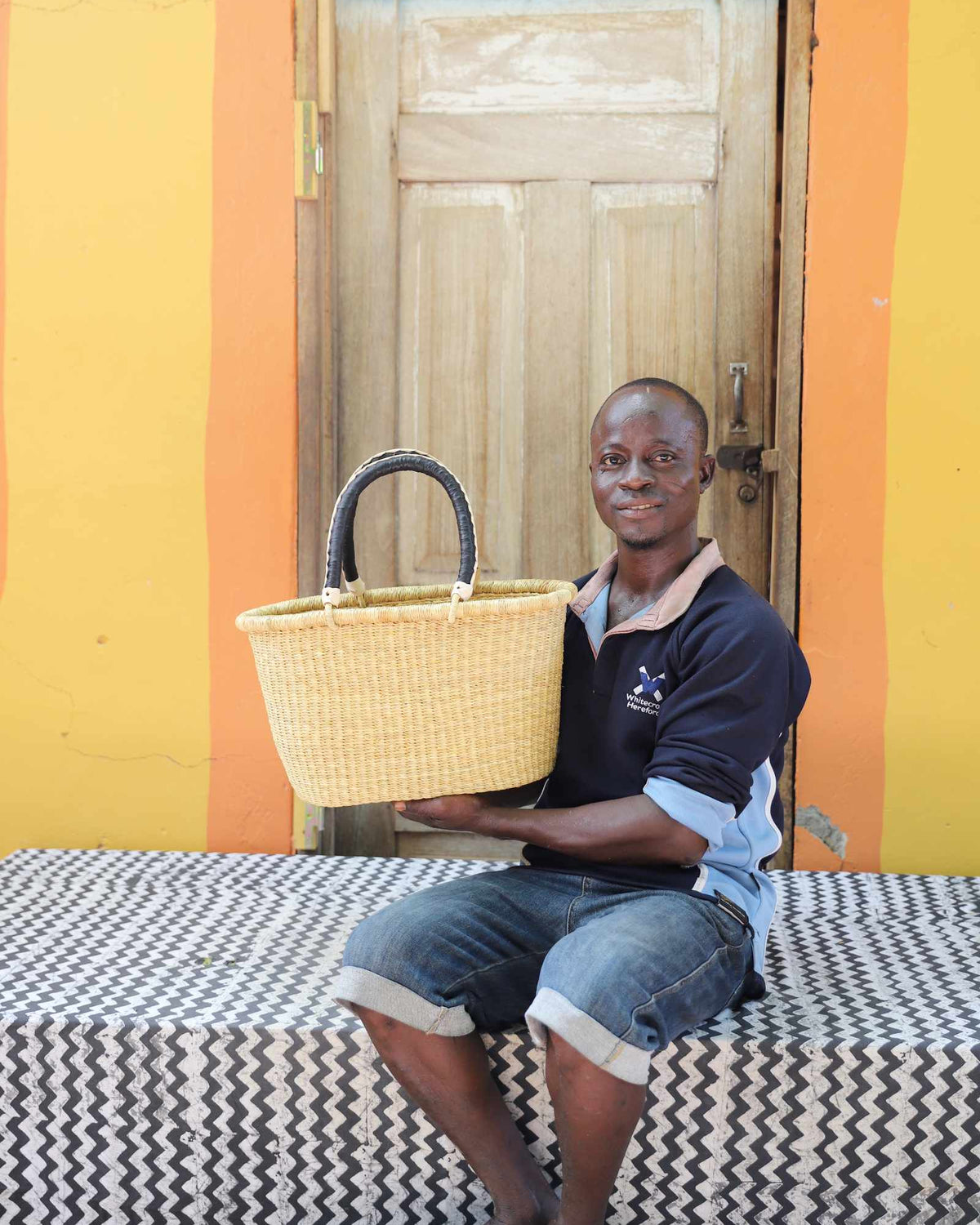 Small Bolga Zaane Tia market basket in plain natural straw, held by a seated man in a dark top.