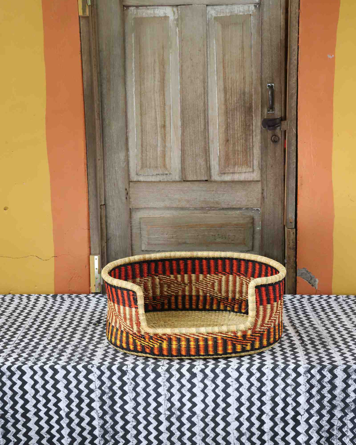 Small handwoven Bolga pet bed in natural straw with bold orange, red, yellow and black vertical stripes.