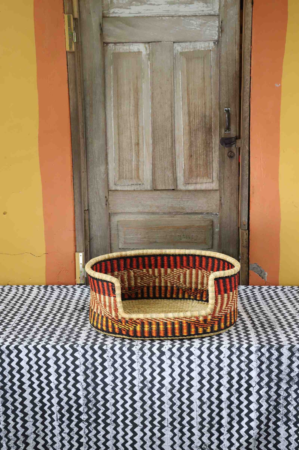 Small handwoven Bolga pet bed in natural straw with bold orange, red, yellow and black vertical stripes.