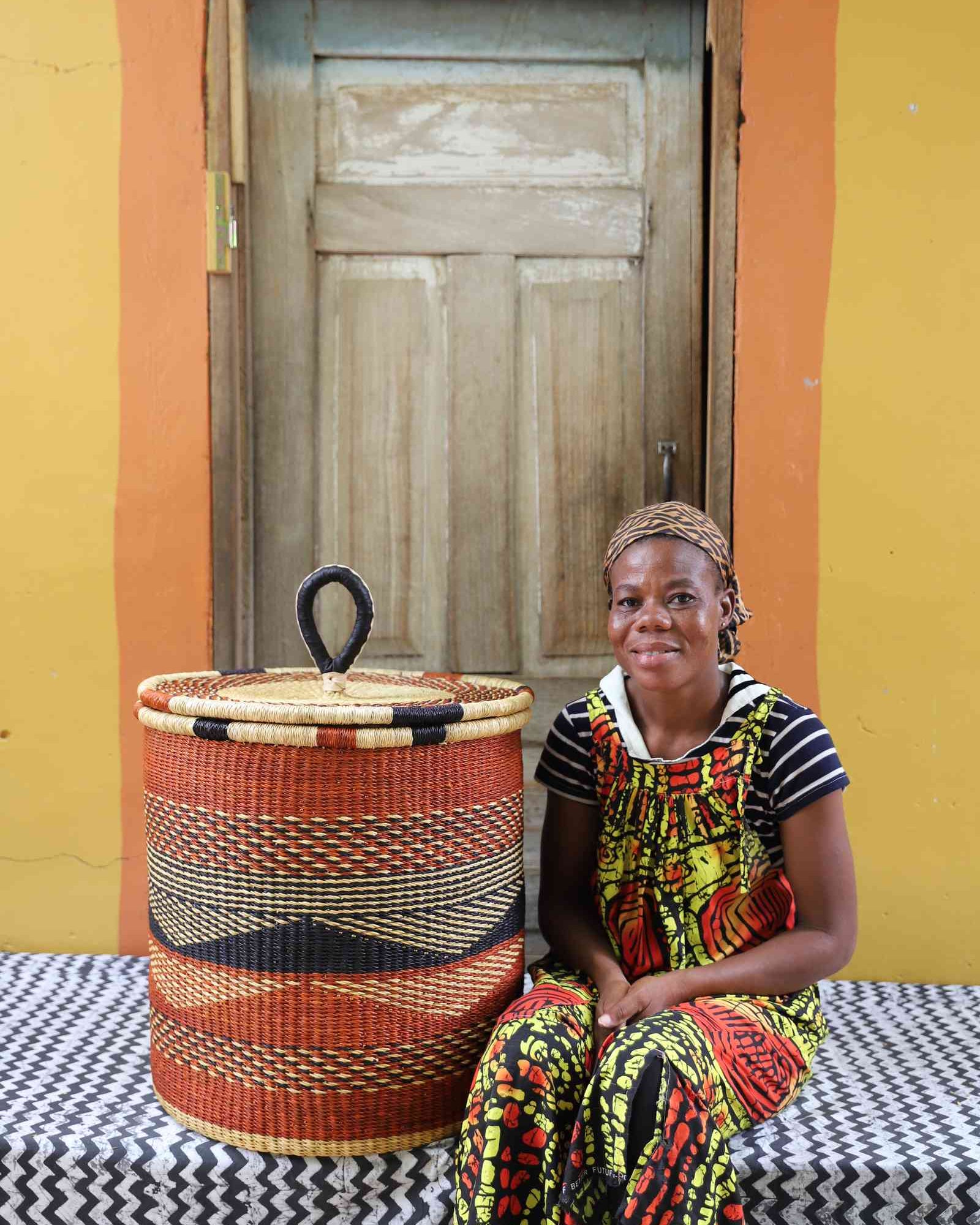 Handwoven Bolga laundry basket in rust-orange with a dark wave motif, styled beside a seated woman.