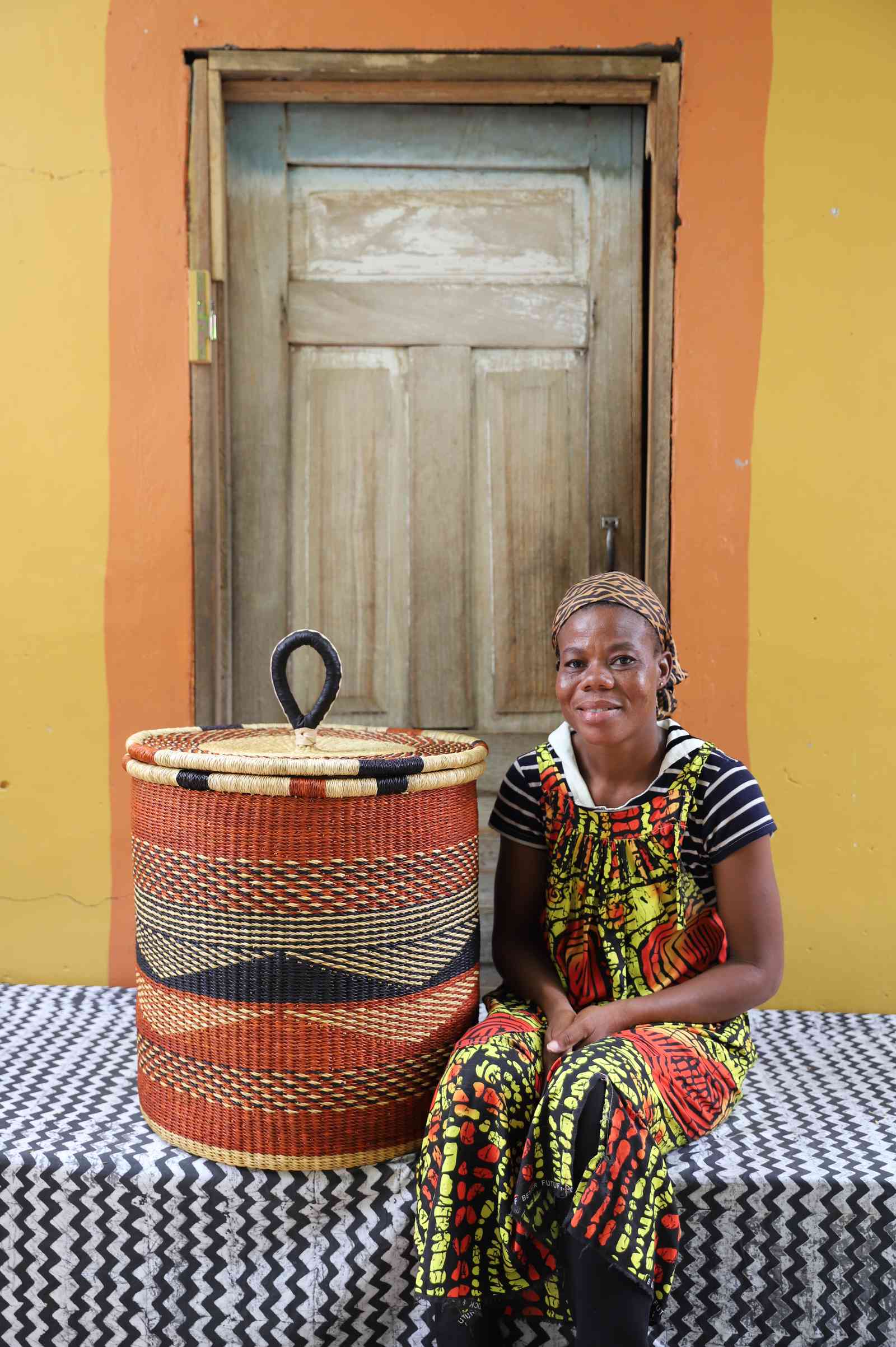 Handwoven Bolga laundry basket in rust-orange with a dark wave motif, styled beside a seated woman.