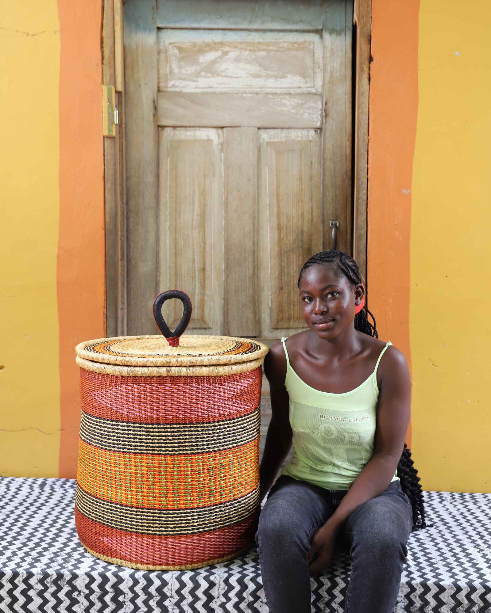 Handwoven Bolga laundry basket in red, orange and yellow bands, styled beside a seated woman in green.