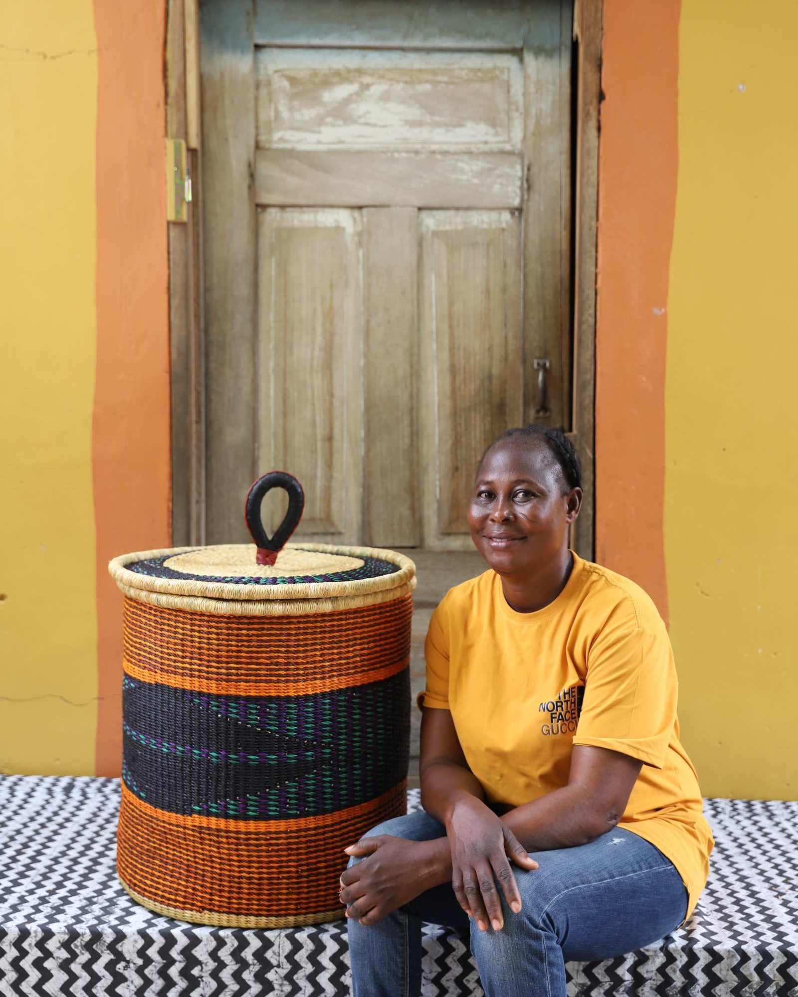 Handwoven Bolga laundry basket with orange and dark blue bands, styled beside a seated woman in yellow.