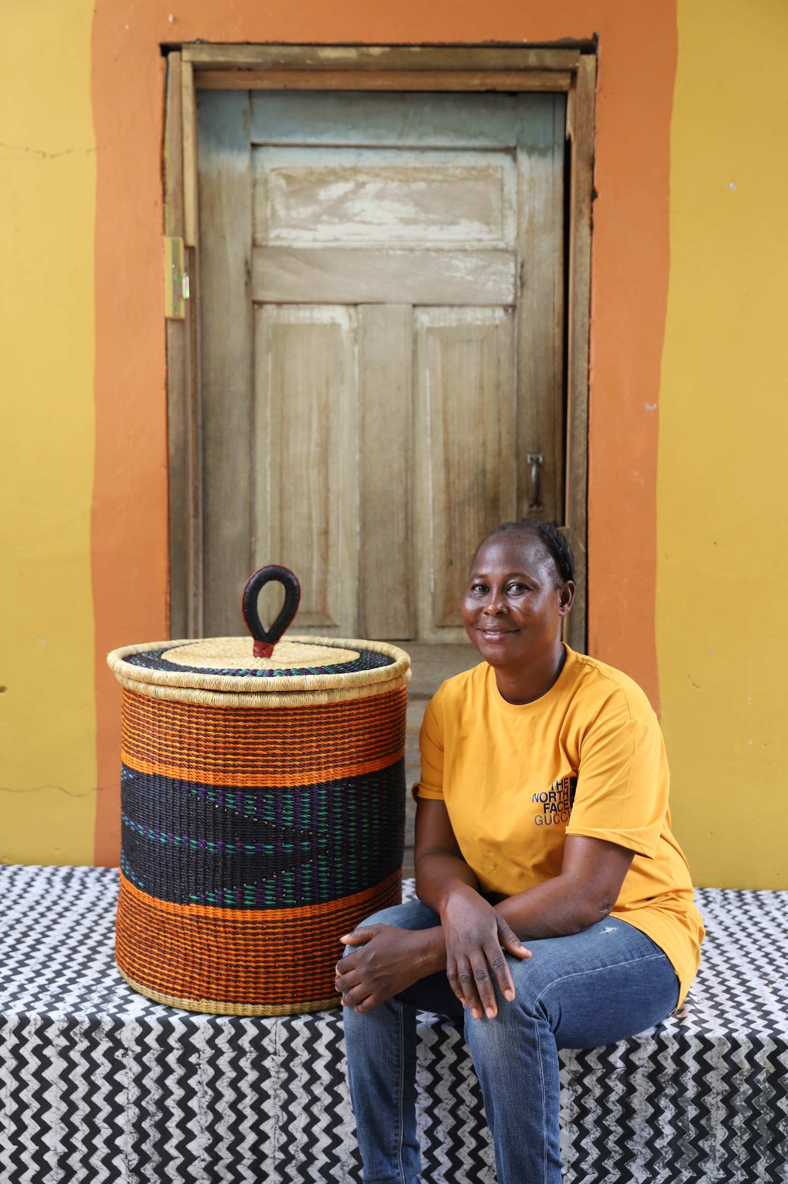 Handwoven Bolga laundry basket with orange and dark blue bands, styled beside a seated woman in yellow.