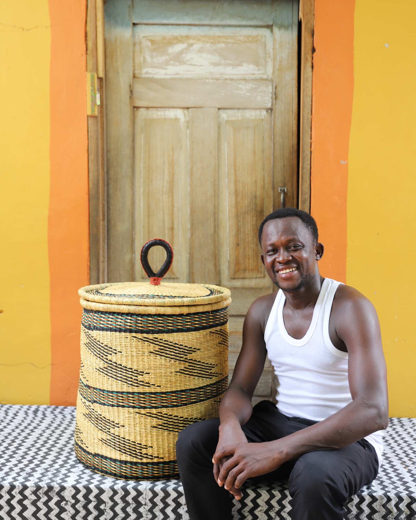 Handwoven Bolga laundry basket in natural straw with black bands, styled beside a seated man in a white vest.