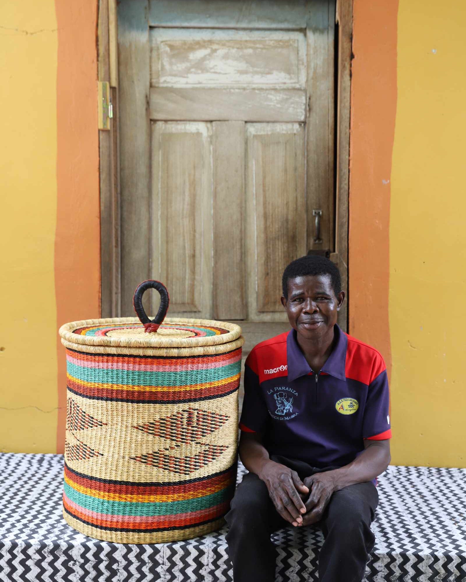 Handwoven Bolga laundry basket in multicolour bands, styled beside a seated man in red and blue.