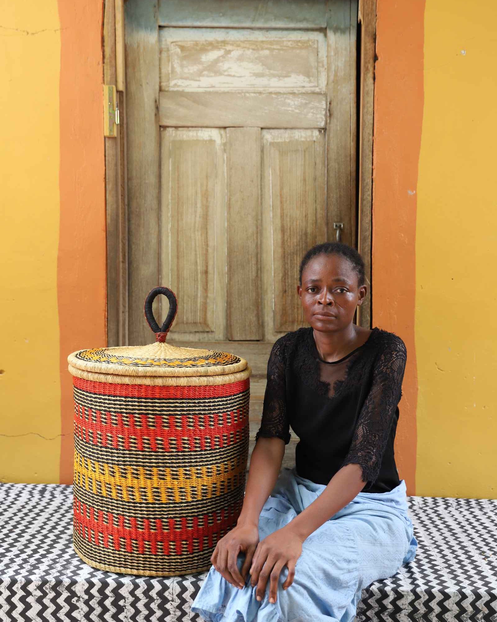 Handwoven Bolga laundry basket in deep red with orange and yellow bands, styled beside a seated woman.