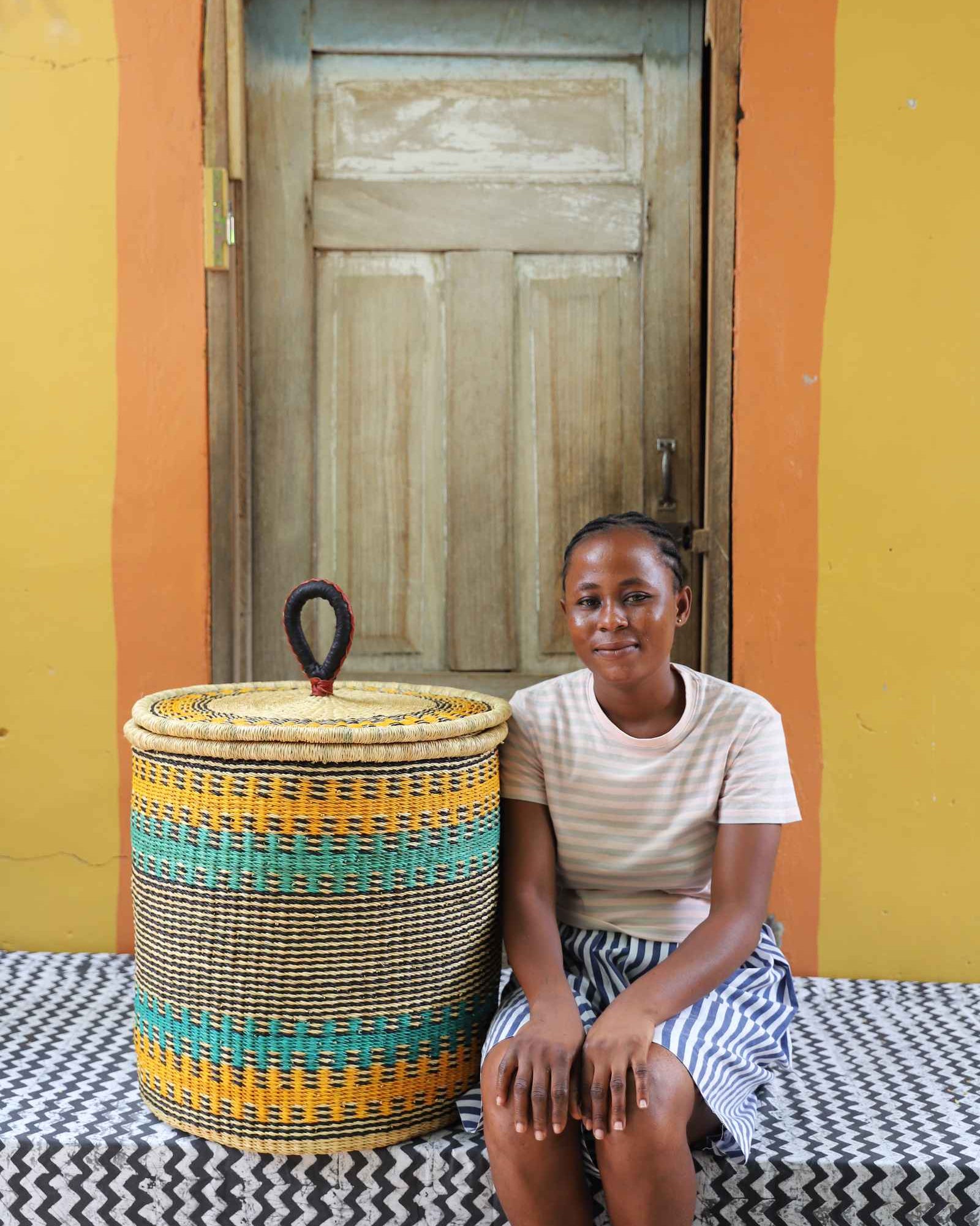 Handwoven Bolga laundry basket striped in yellow-orange and teal, styled beside a seated woman in stripes.