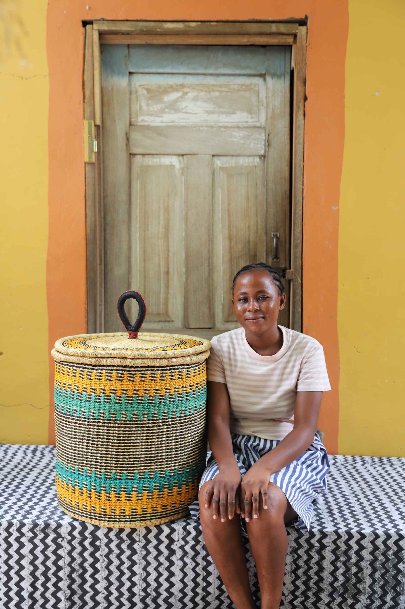 Handwoven Bolga laundry basket striped in yellow-orange and teal, styled beside a seated woman in stripes.