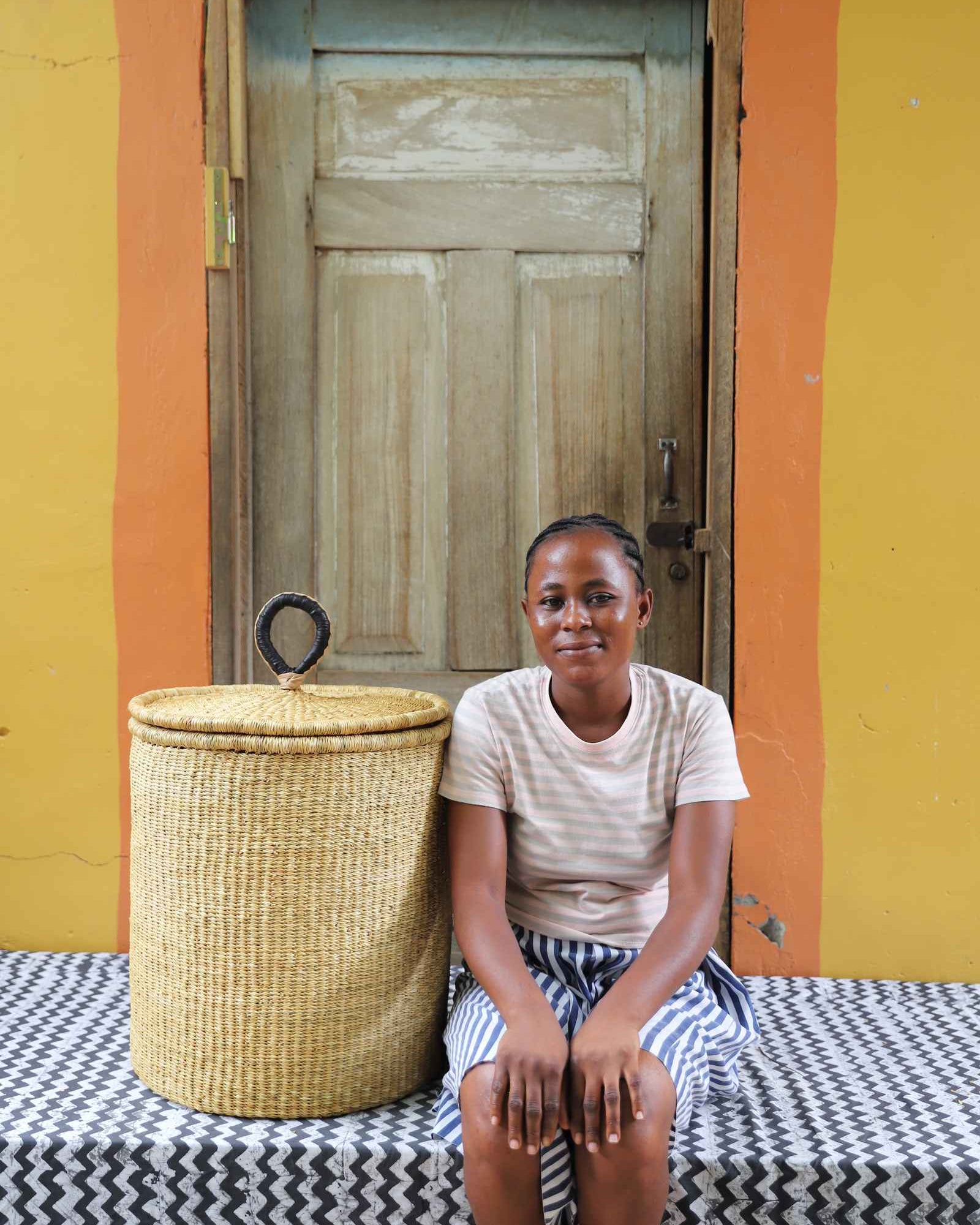 Handwoven Bolga laundry basket with coiled rim and lid in natural elephant grass, styled beside a seated woman.