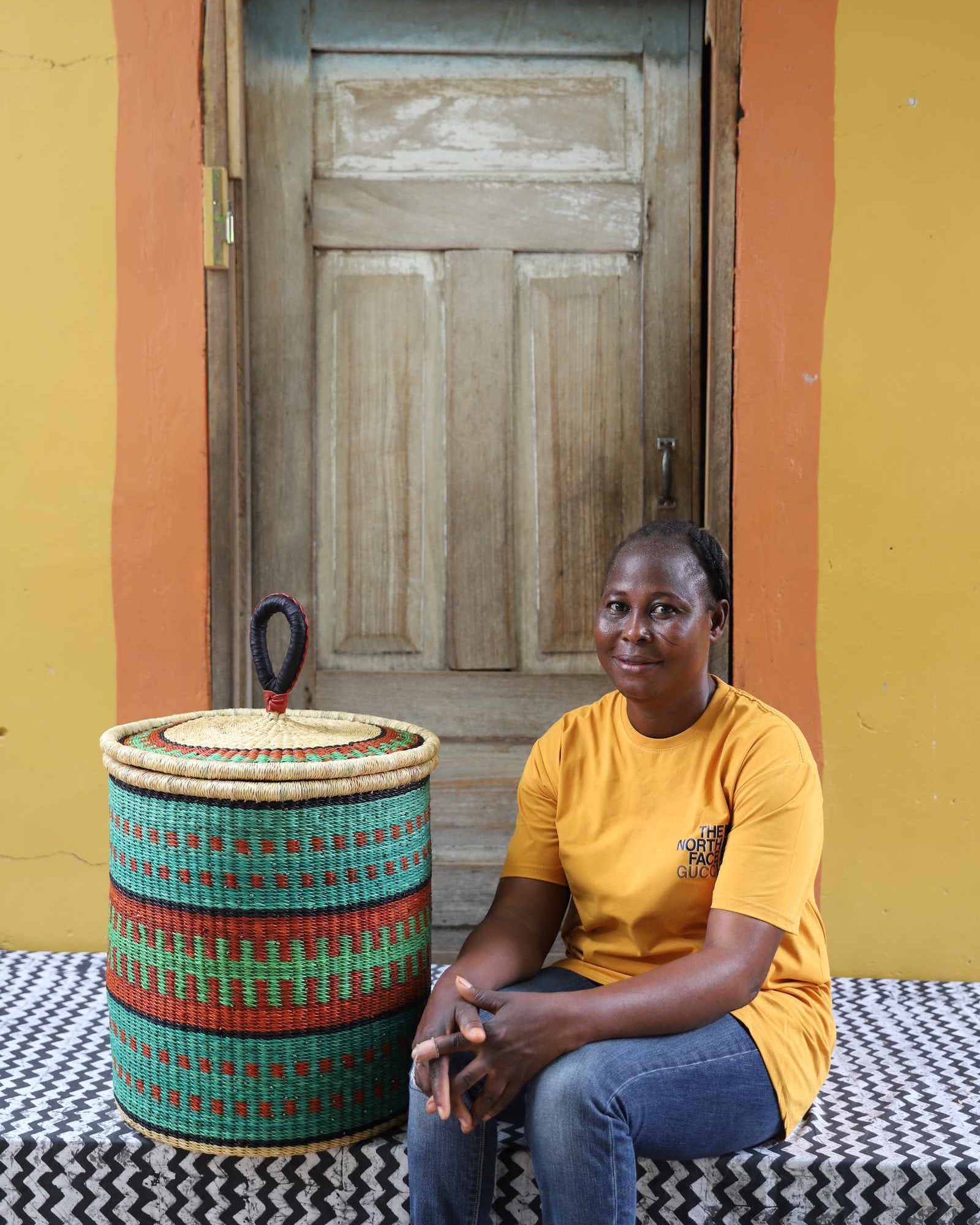Handwoven Bolga laundry basket in teal, red and green bands, styled beside a seated woman in yellow.