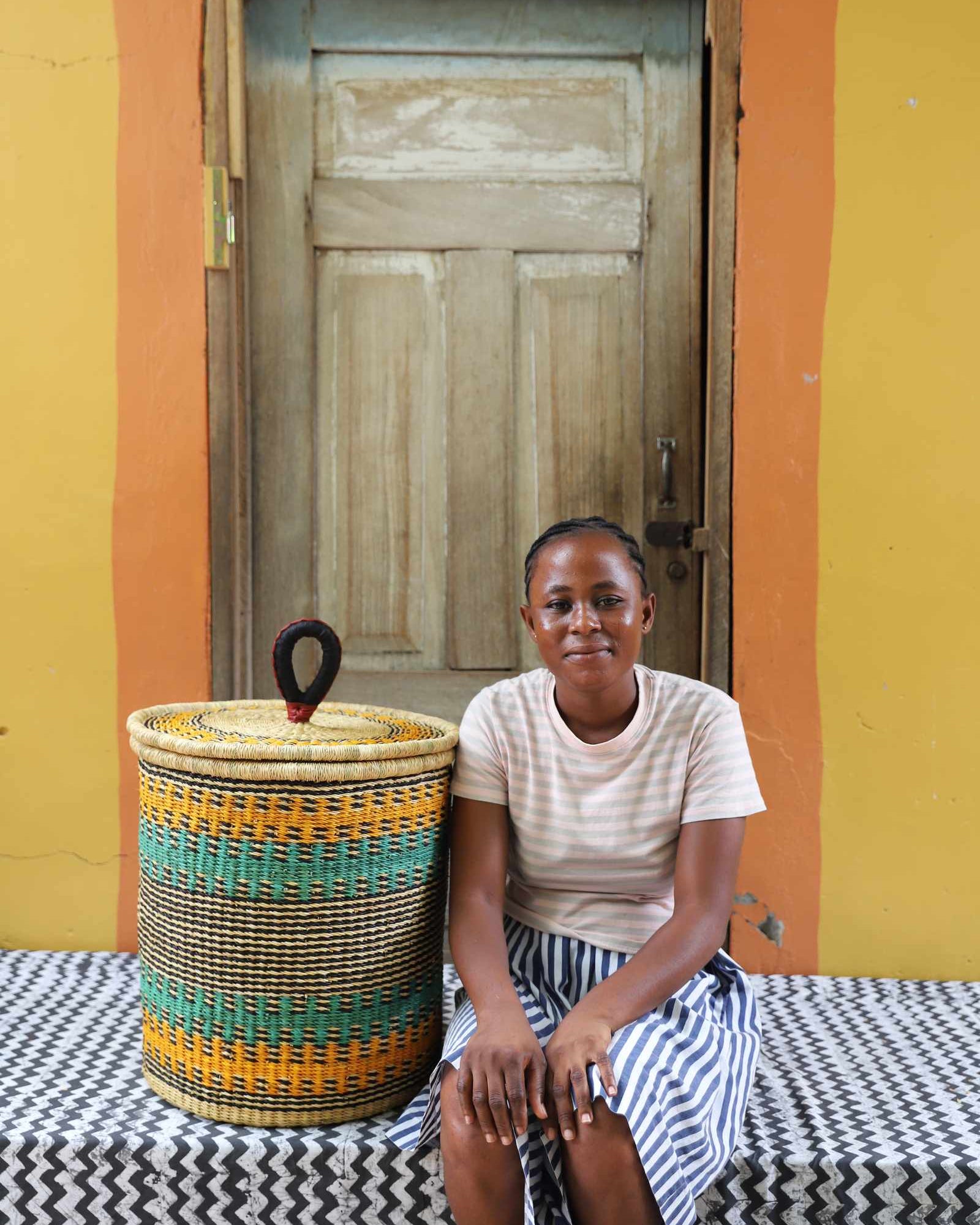 Handwoven Bolga laundry basket in natural straw with orange and teal bands, styled beside a seated woman.