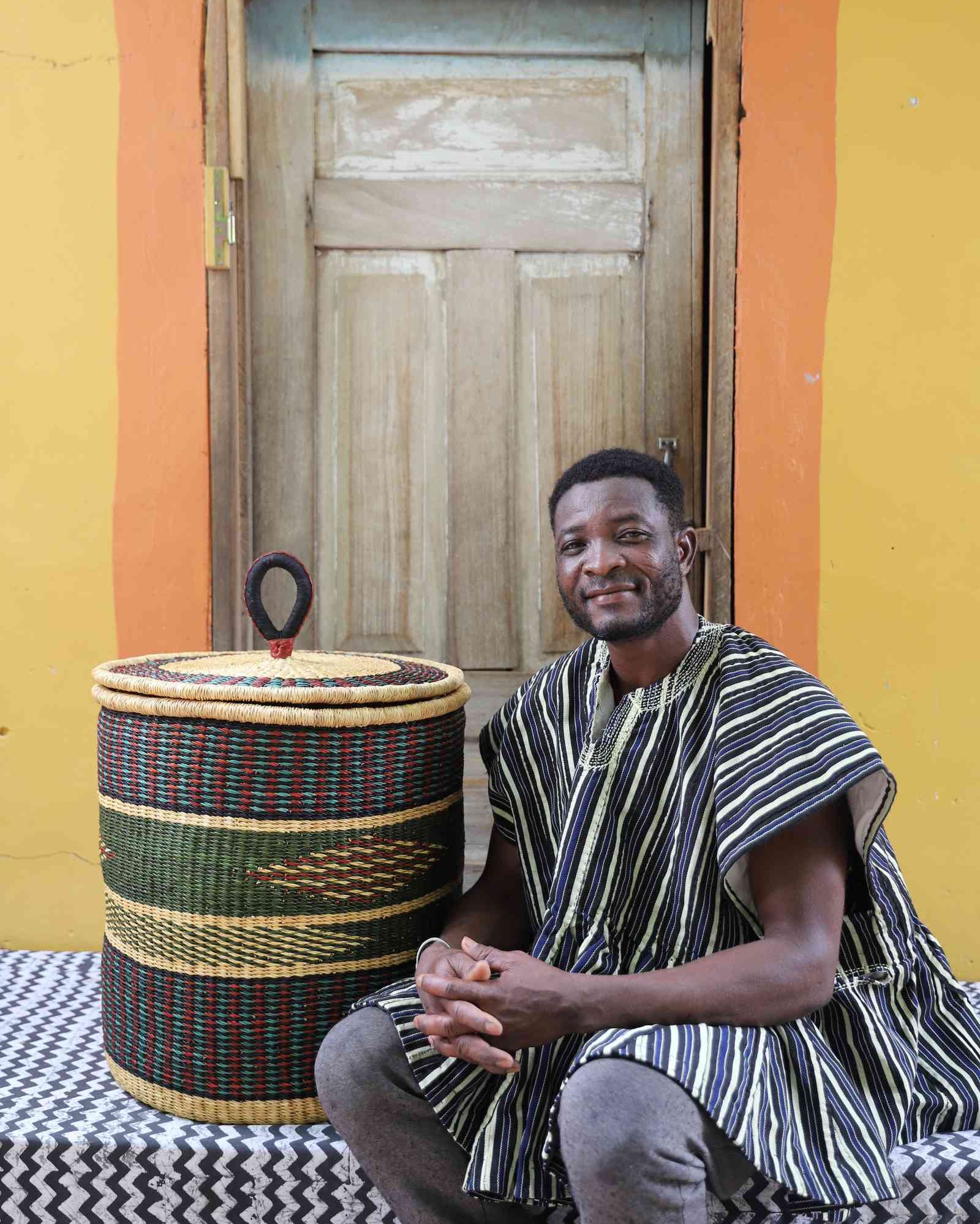 Handwoven Bolga laundry basket in natural straw with a dark green band, styled beside a seated man in stripes.
