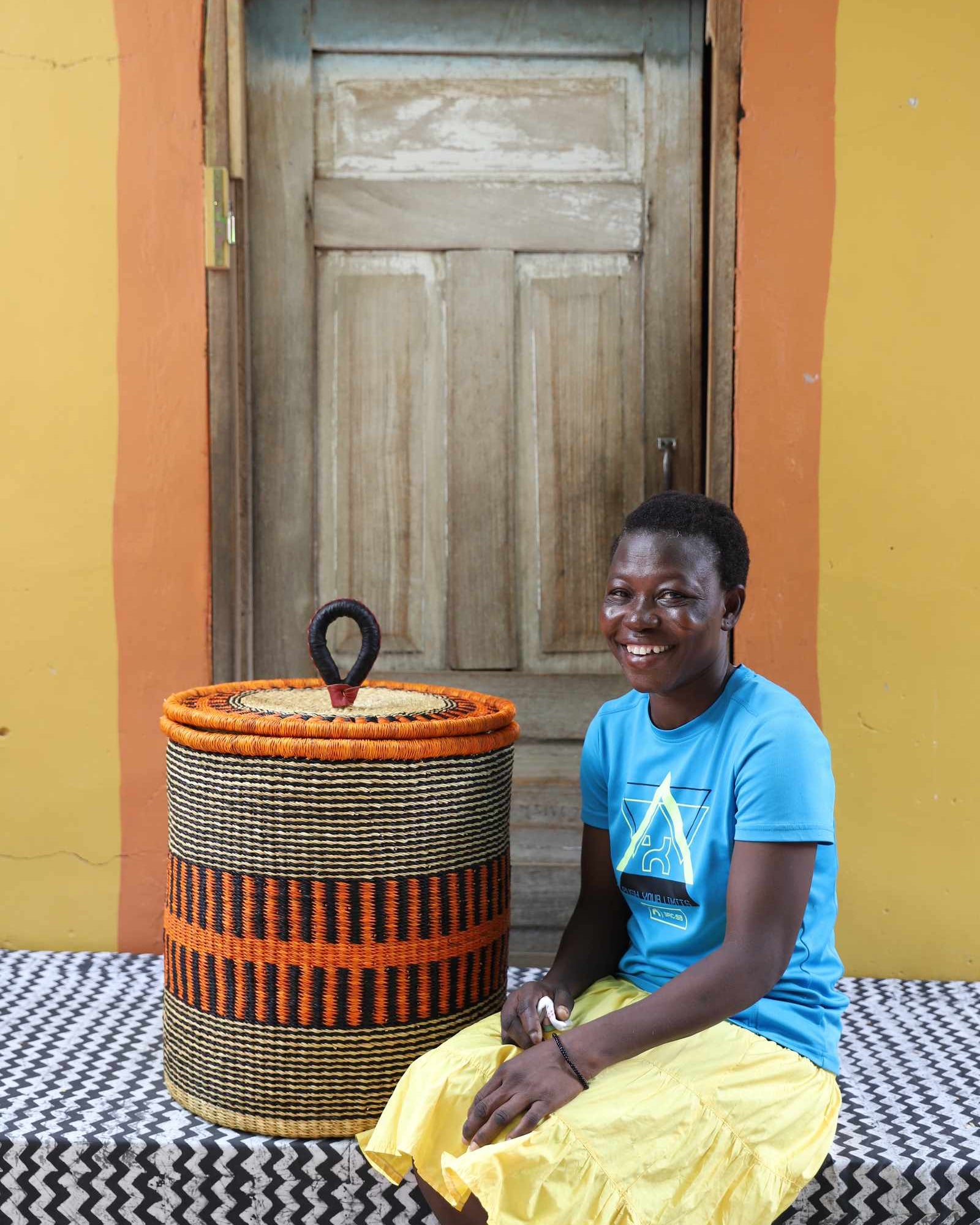 Handwoven Bolga laundry basket in natural straw with an orange band, styled beside a seated woman in yellow.