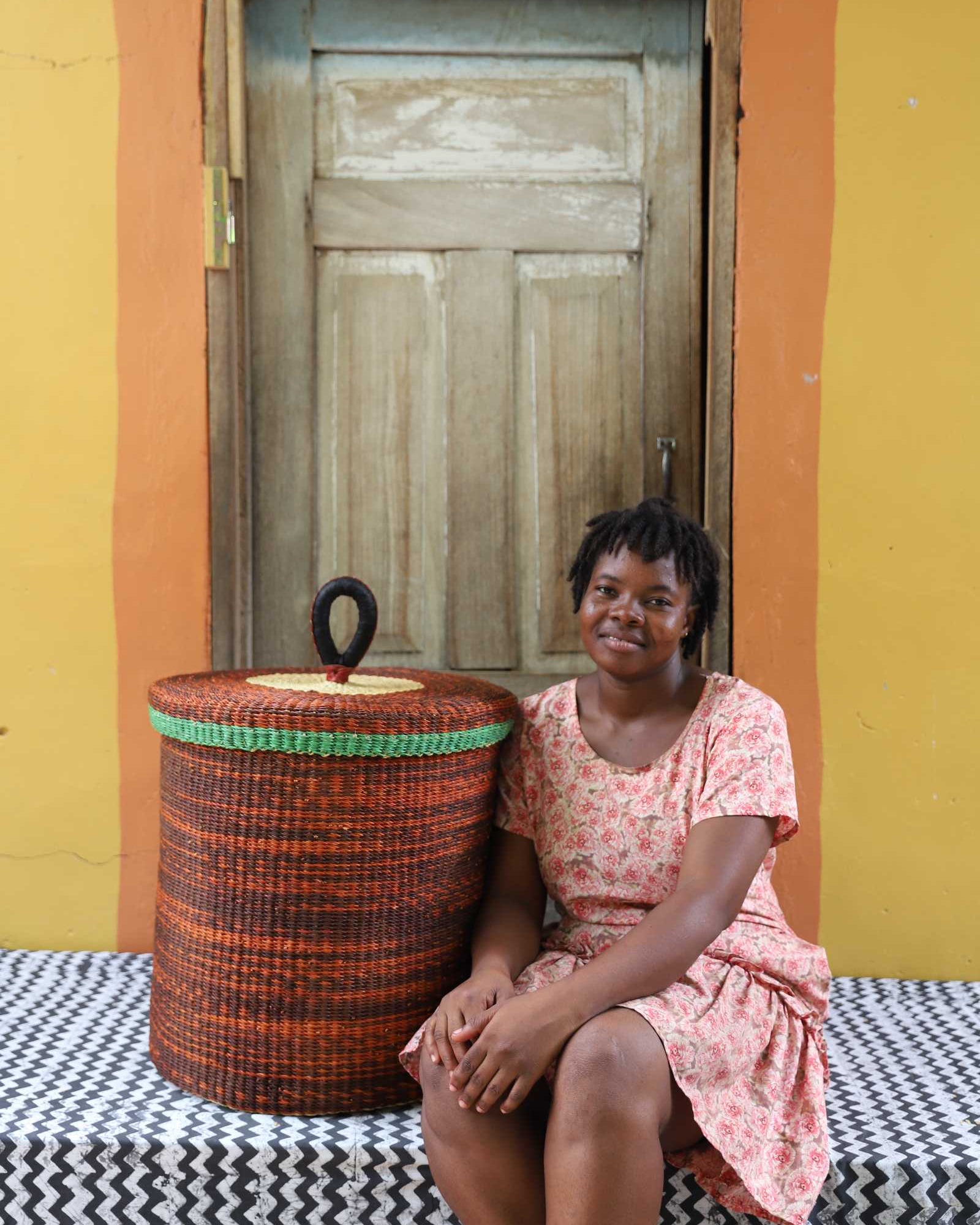 Handwoven Bolga laundry basket with lid in rust-orange with a green rim, styled beside a seated woman.