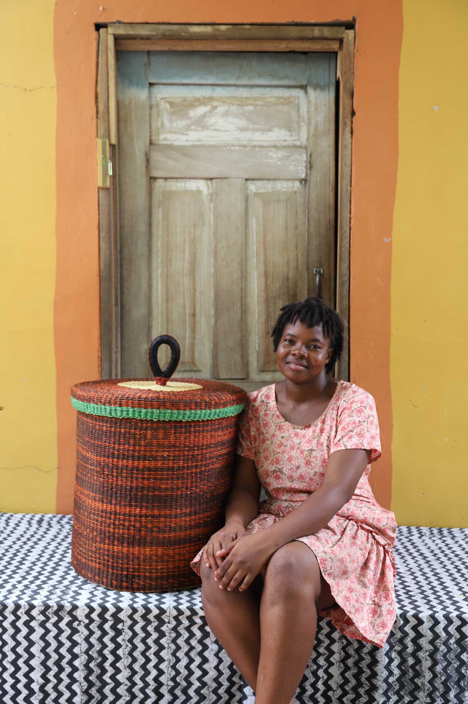 Handwoven Bolga laundry basket with lid in rust-orange with a green rim, styled beside a seated woman.