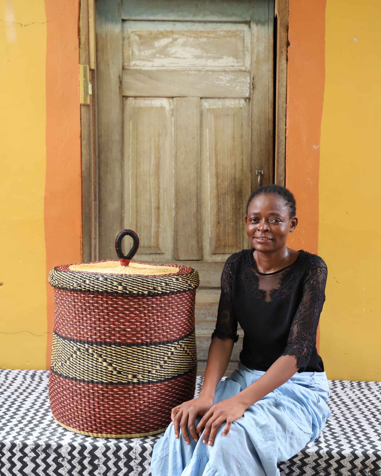 Handwoven Bolga laundry basket with lid in deep red with a black accent band, styled beside a seated woman.