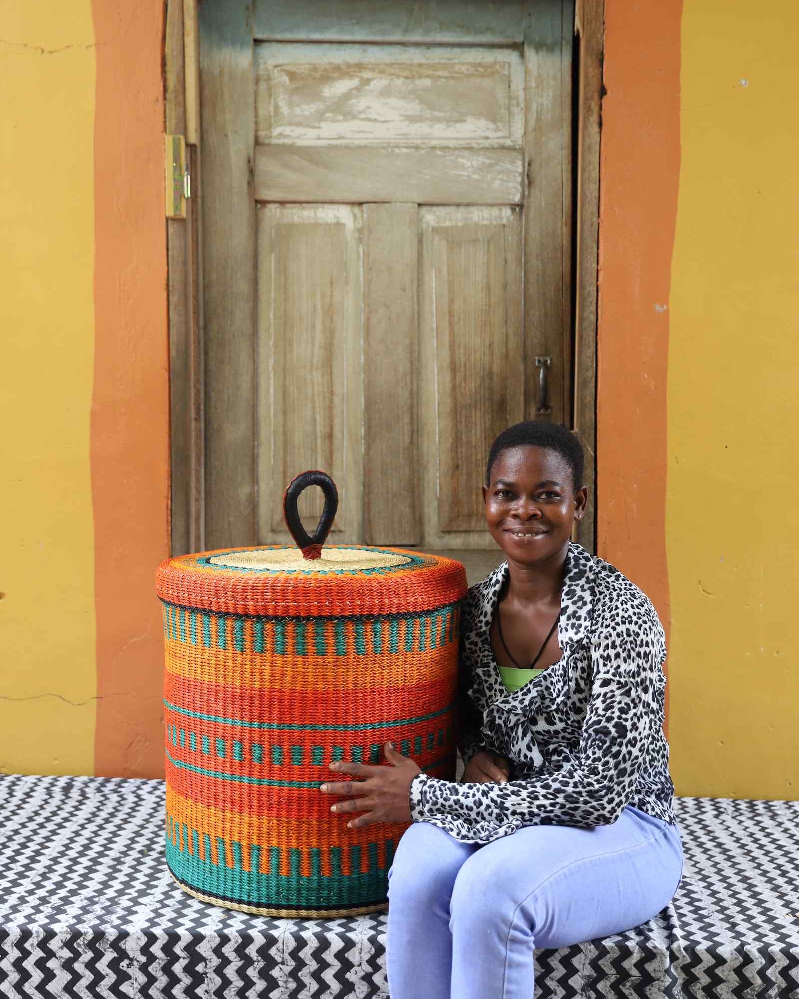 Handwoven Bolga laundry basket with lid in orange and teal, styled beside a seated woman.