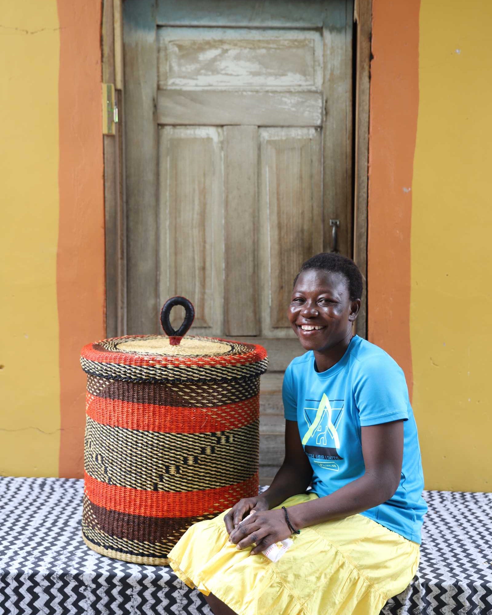 Handwoven Bolga laundry basket with lid in red and black bands, styled beside a seated woman in yellow.