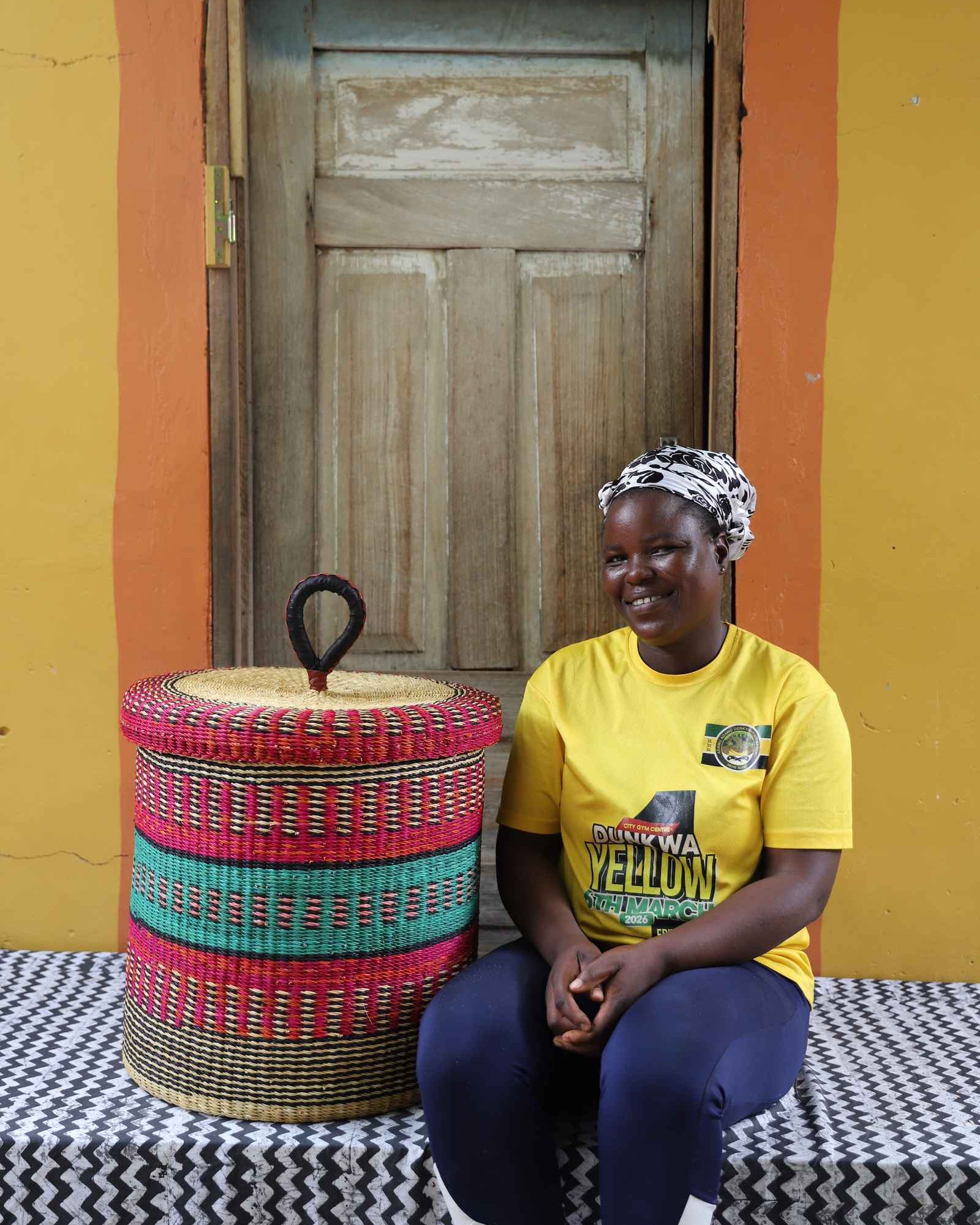 Handwoven Bolga laundry basket with lid in red and green bands, styled beside a seated woman in a yellow top.